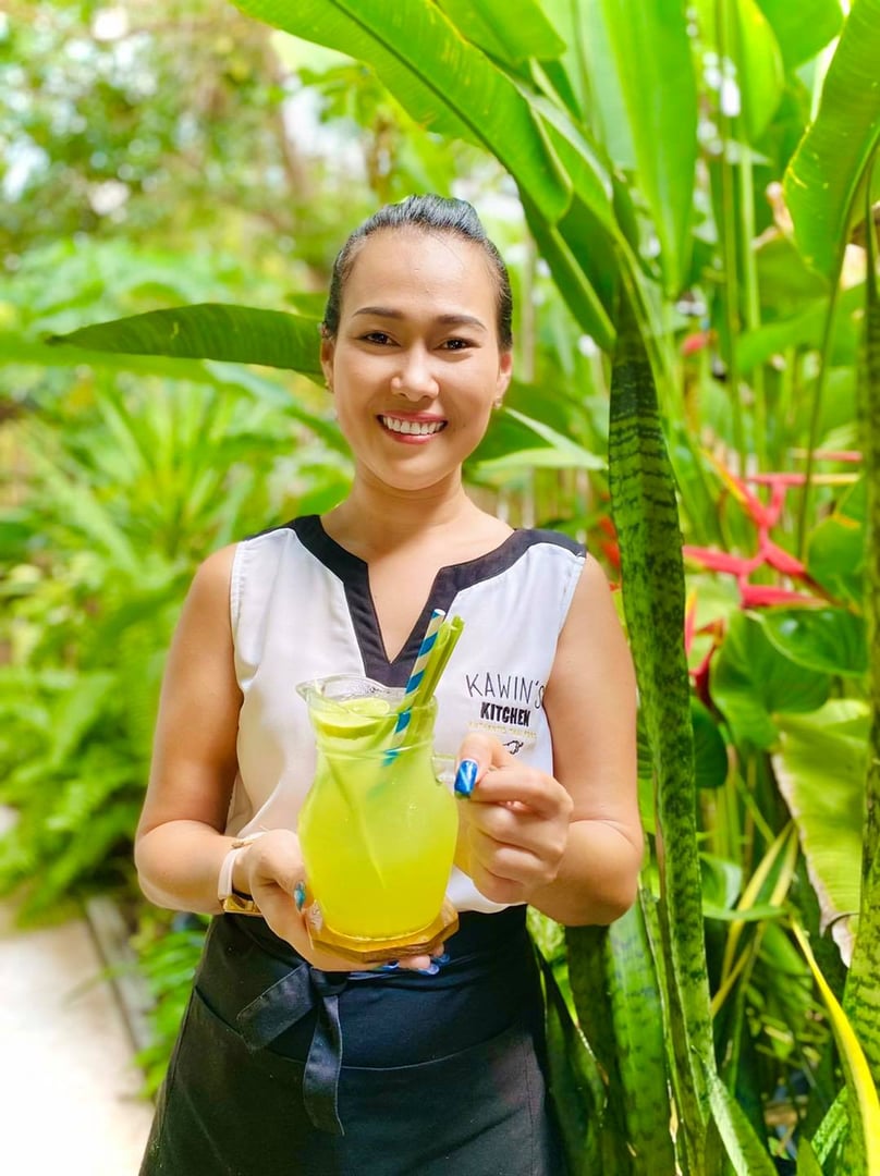 Smiling woman holding a pitcher of yellow juice with a straw, standing in a lush green garden.