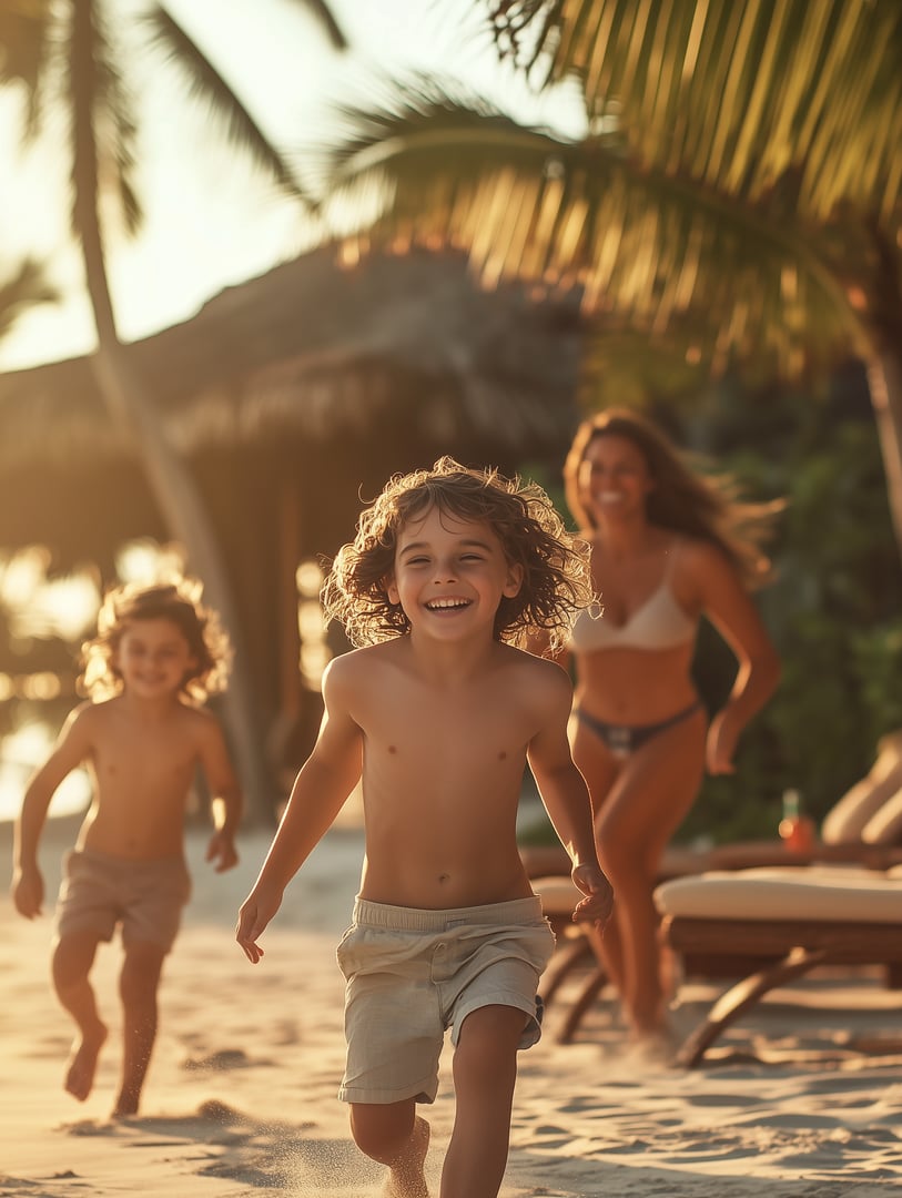 Two kids joyfully running on a sunny beach, with a woman following behind under palm trees and a thatched hut.