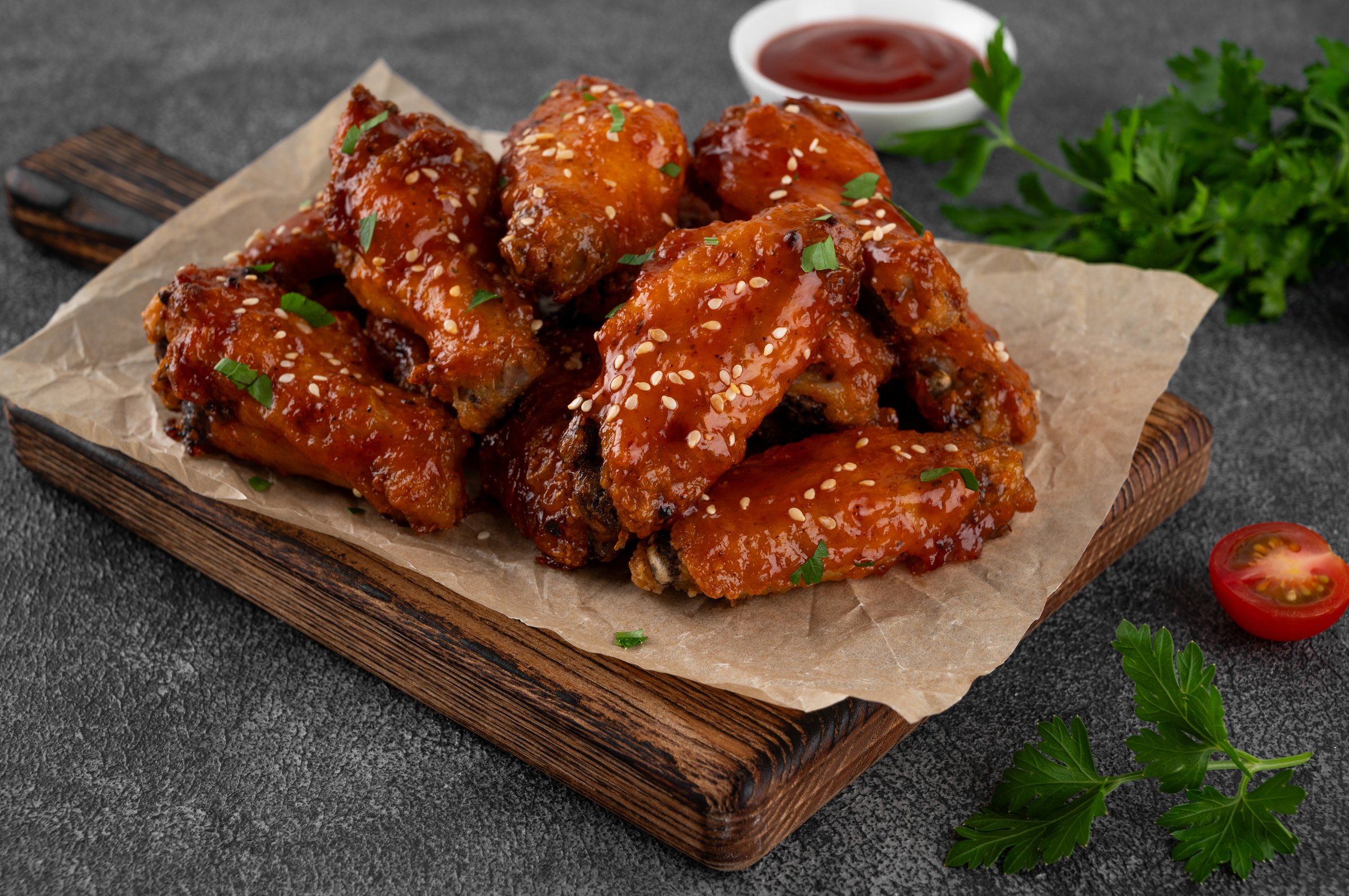 Baked chicken wings in a sweet chili sauce and sesame seeds on a wooden board on a dark concrete background