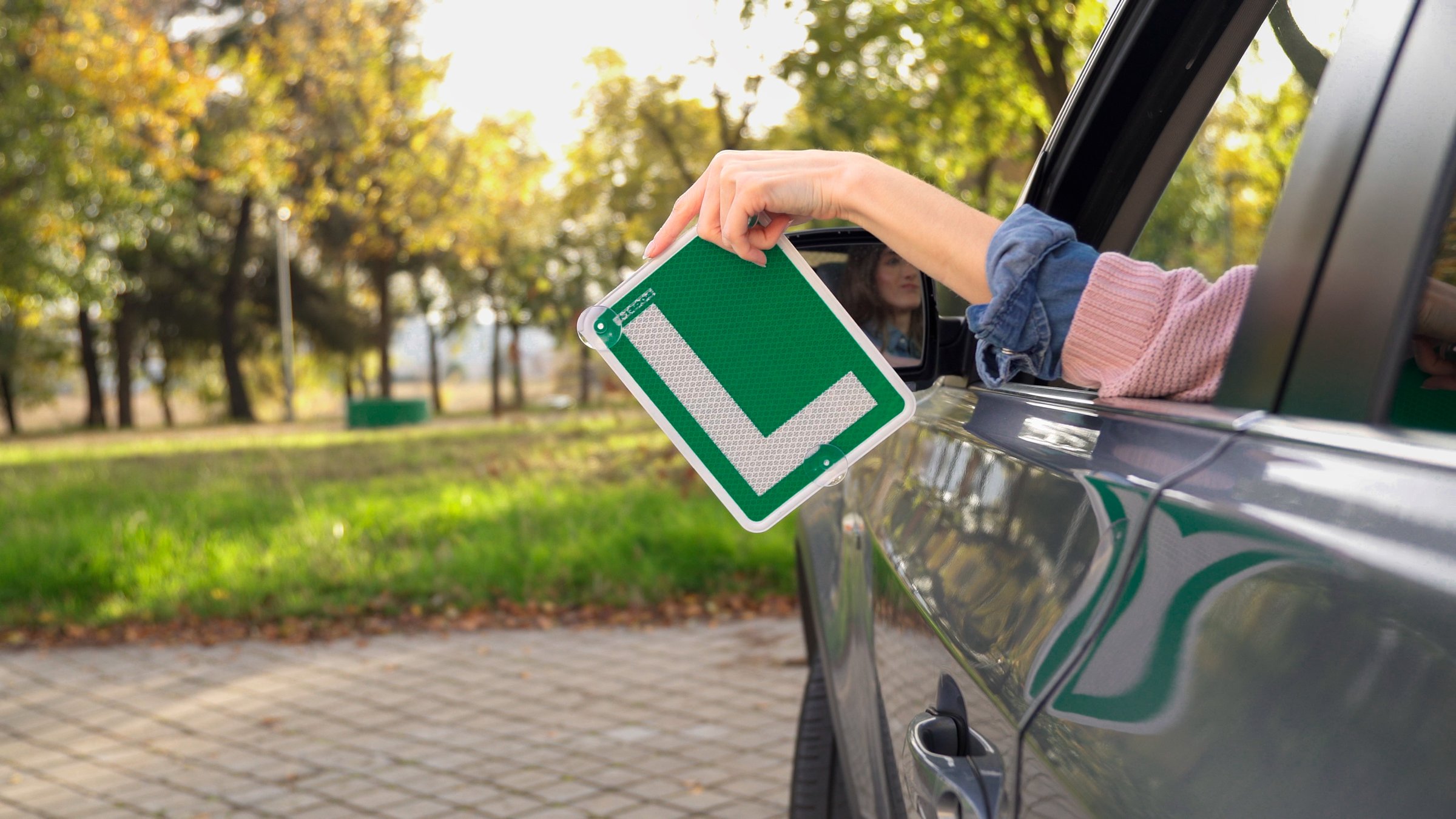 Young woman driving holding a learner l plate out of the car window, celebrating passing her driving test