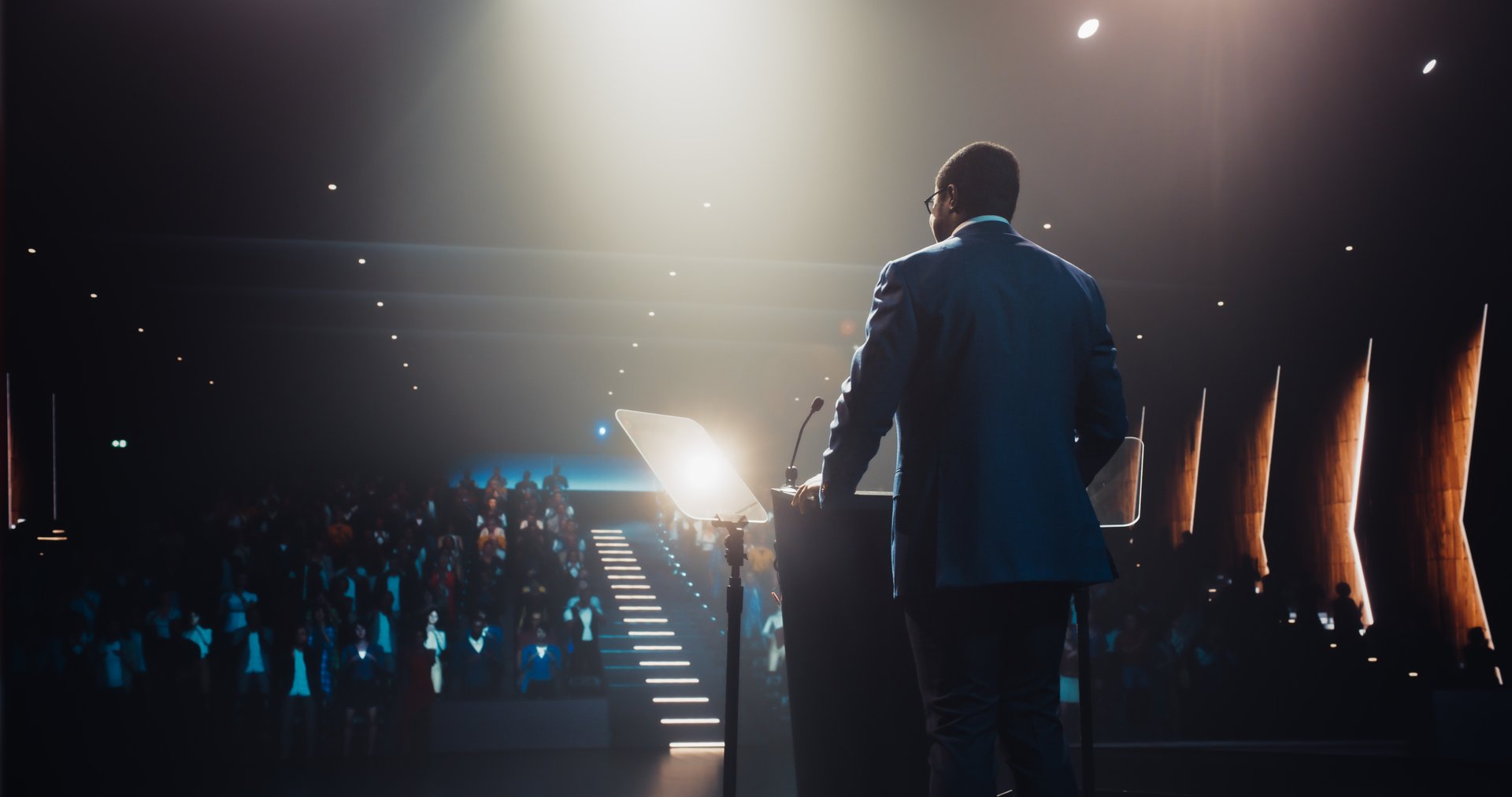 Confident Thought Leader Walking to a Tribune with laptop Computer, Delivering an Inspiring Presentation About Innovation at Technology Conference. Black Man Talking to an Audience in a Dark Venue