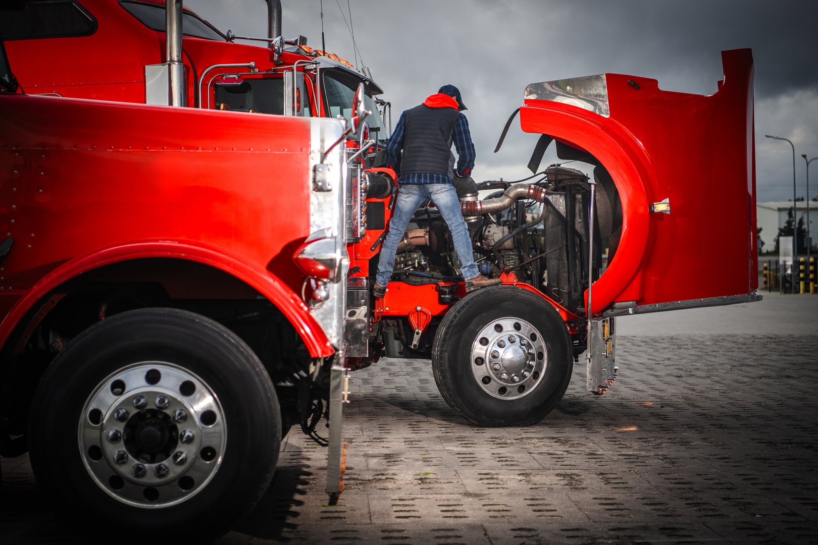 A mechanic works on the engine of a red truck at a transportation center under cloudy skies during the afternoon.