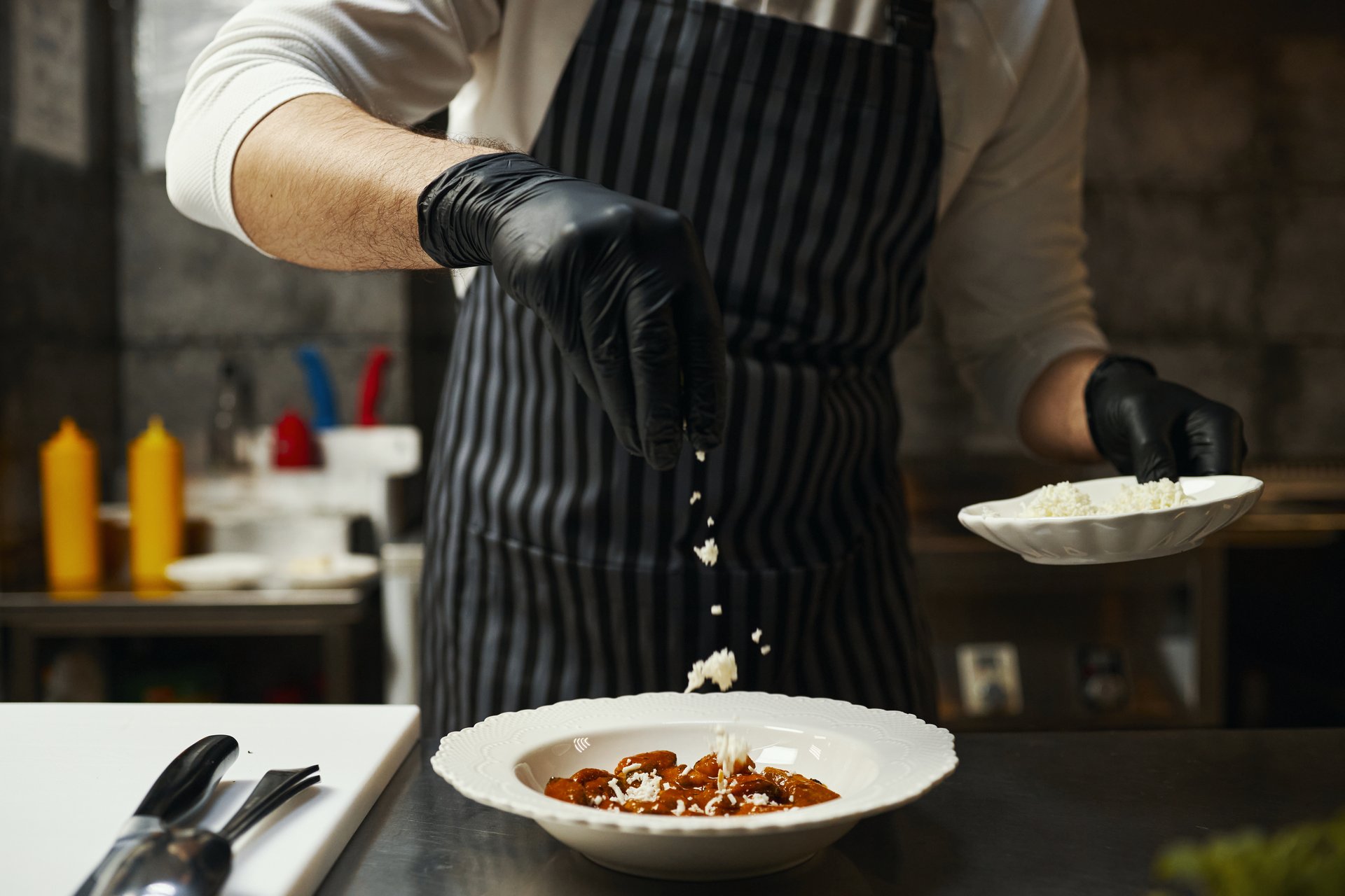 Caucasian young adult man wearing striped apron and black gloves garnishing prepared dish with crumbled cheese in professional kitchen, holding plate in one hand while sprinkling topping