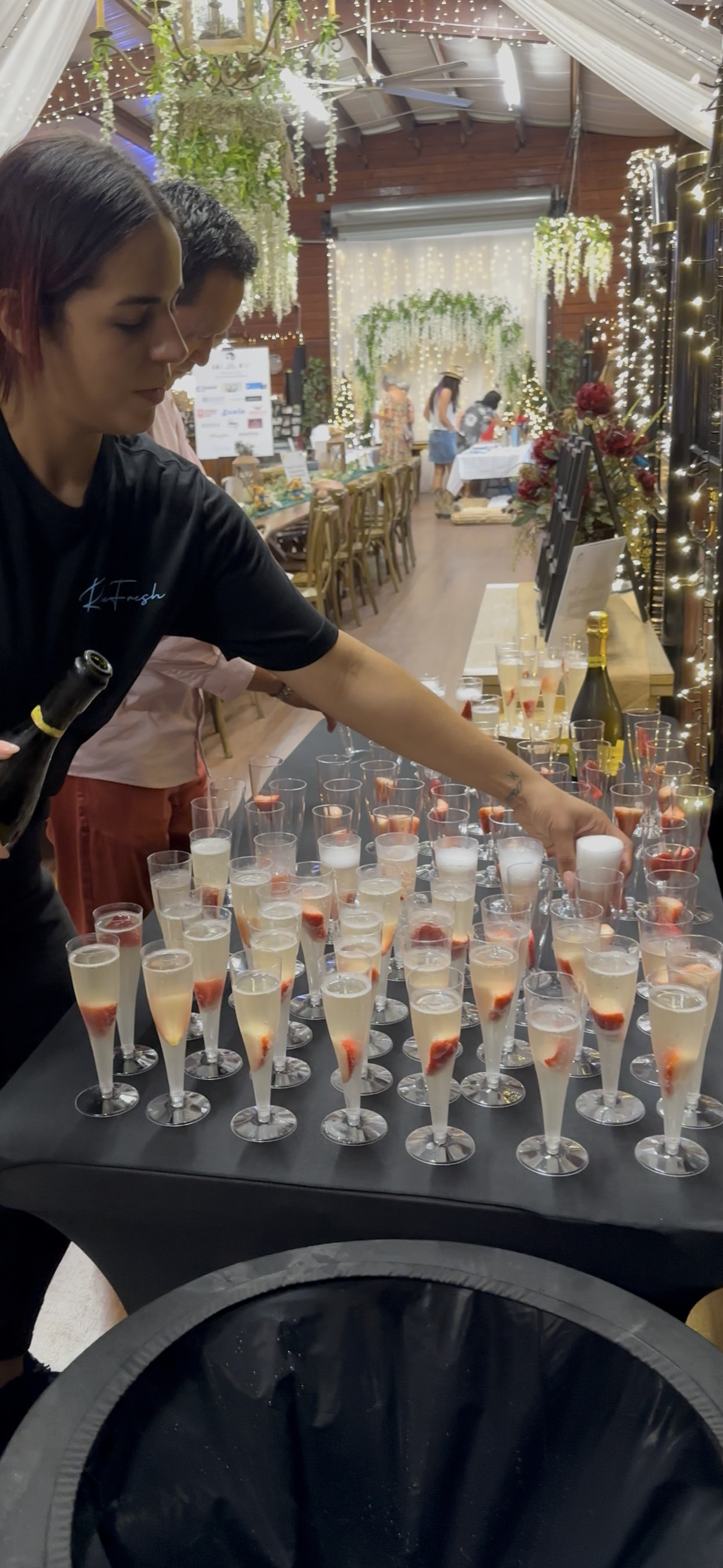 A server arranging champagne glasses with strawberries on a black table at a decorated event venue.