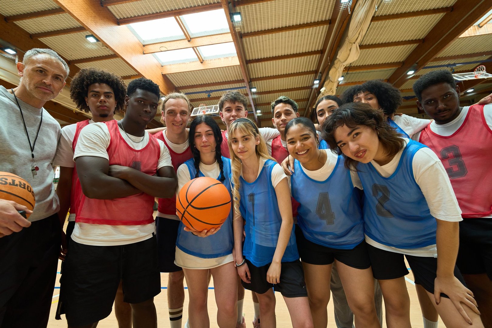 High school basketball team of diverse teenagers and their coach standing together for a group photo, holding a basketball, in a gymnasium