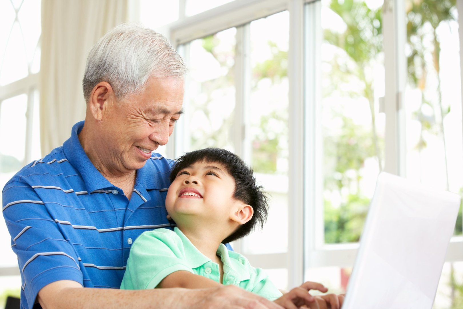 Chinese Grandfather And Grandson Sitting At Desk Using Laptop At Home Smiling At Each Other