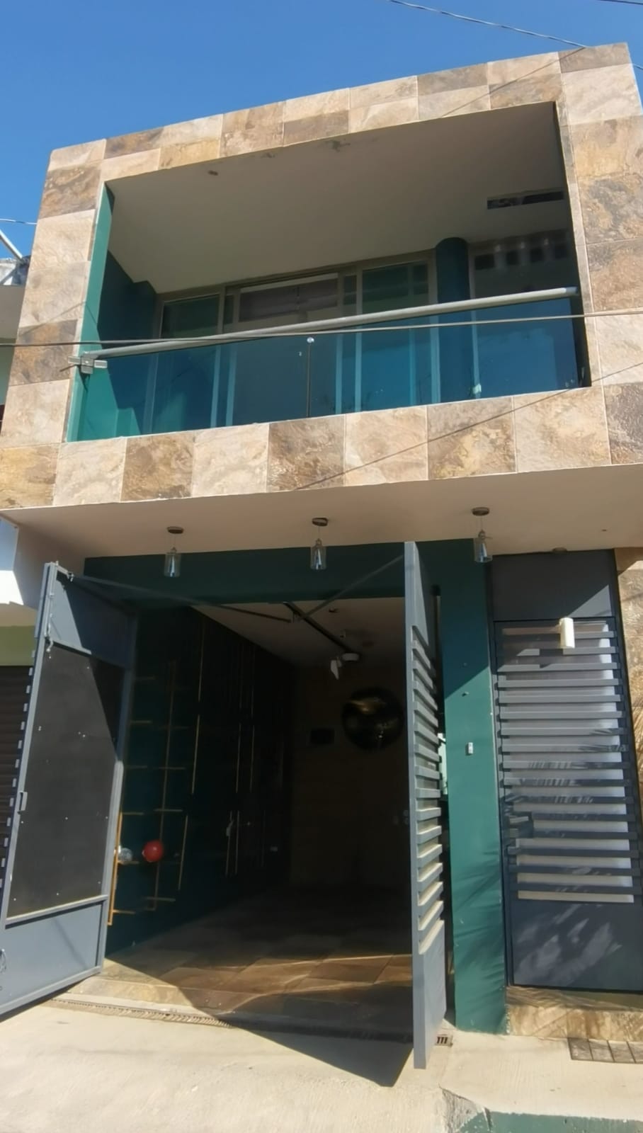 Modern two-story house with stone facade, glass balcony, and open metal gate on a sunny day.