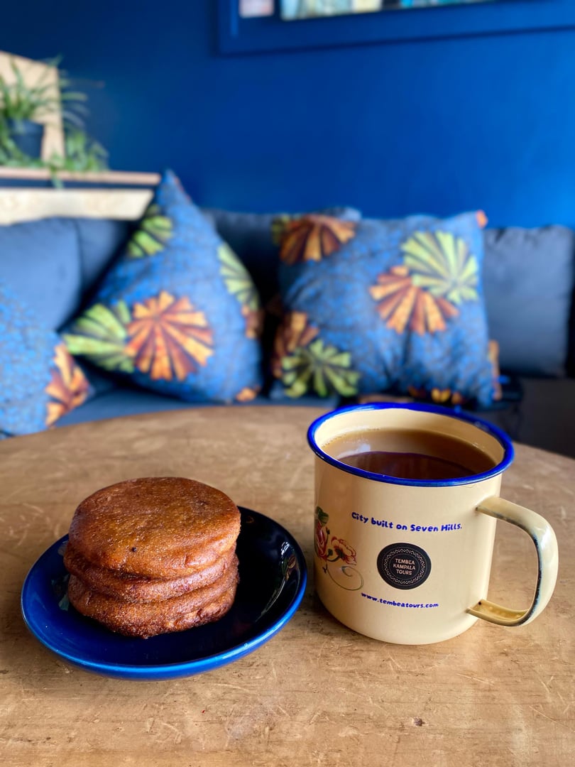 A stack of brown pancakes on a blue plate next to a mug of tea on a wooden table, with a blue couch and pillows in the background.