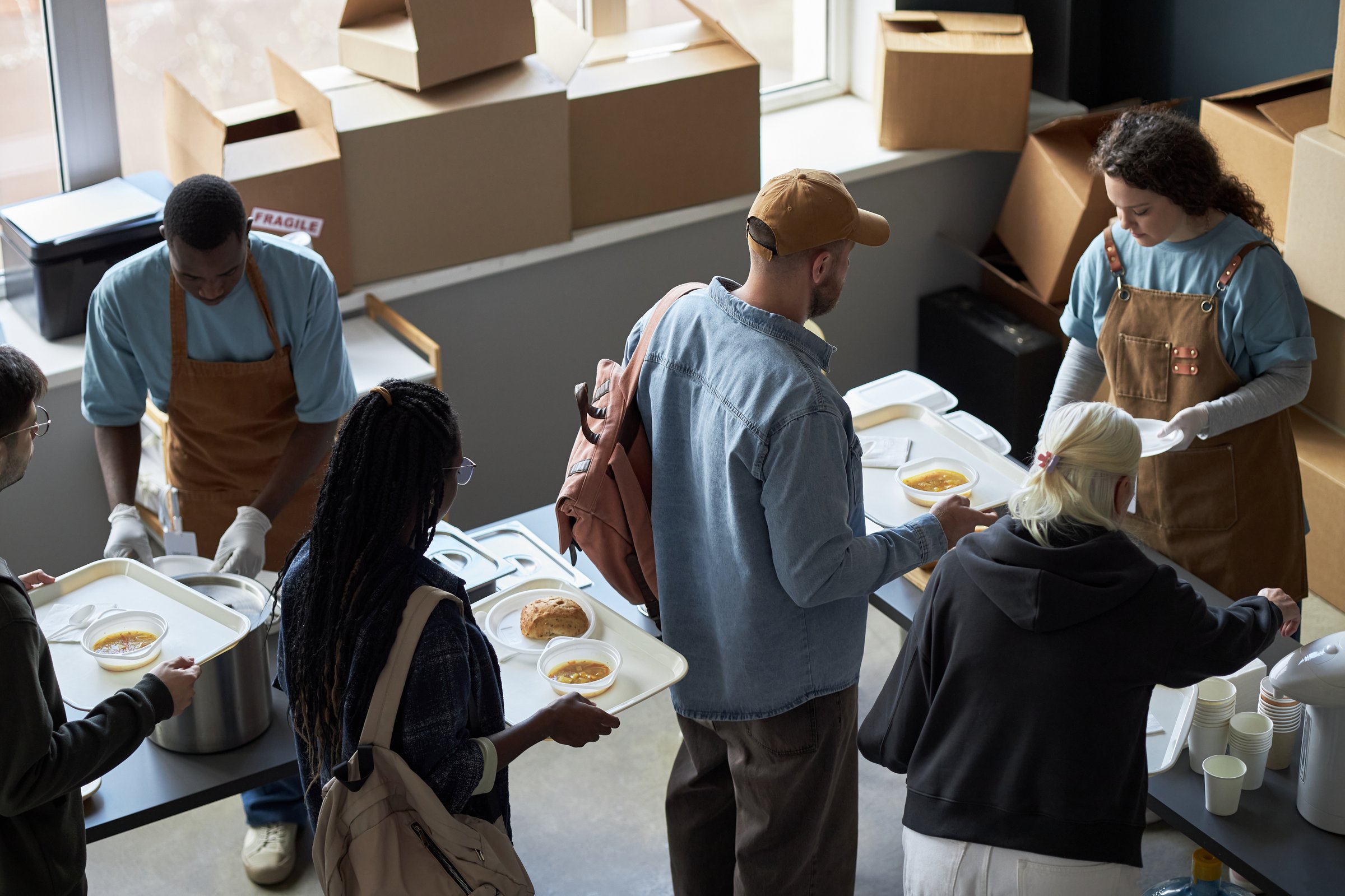Diverse group of individuals engaged in serving food at a community center. Volunteers handling trays and preparing meals in a well-lit room, showcasing team effort