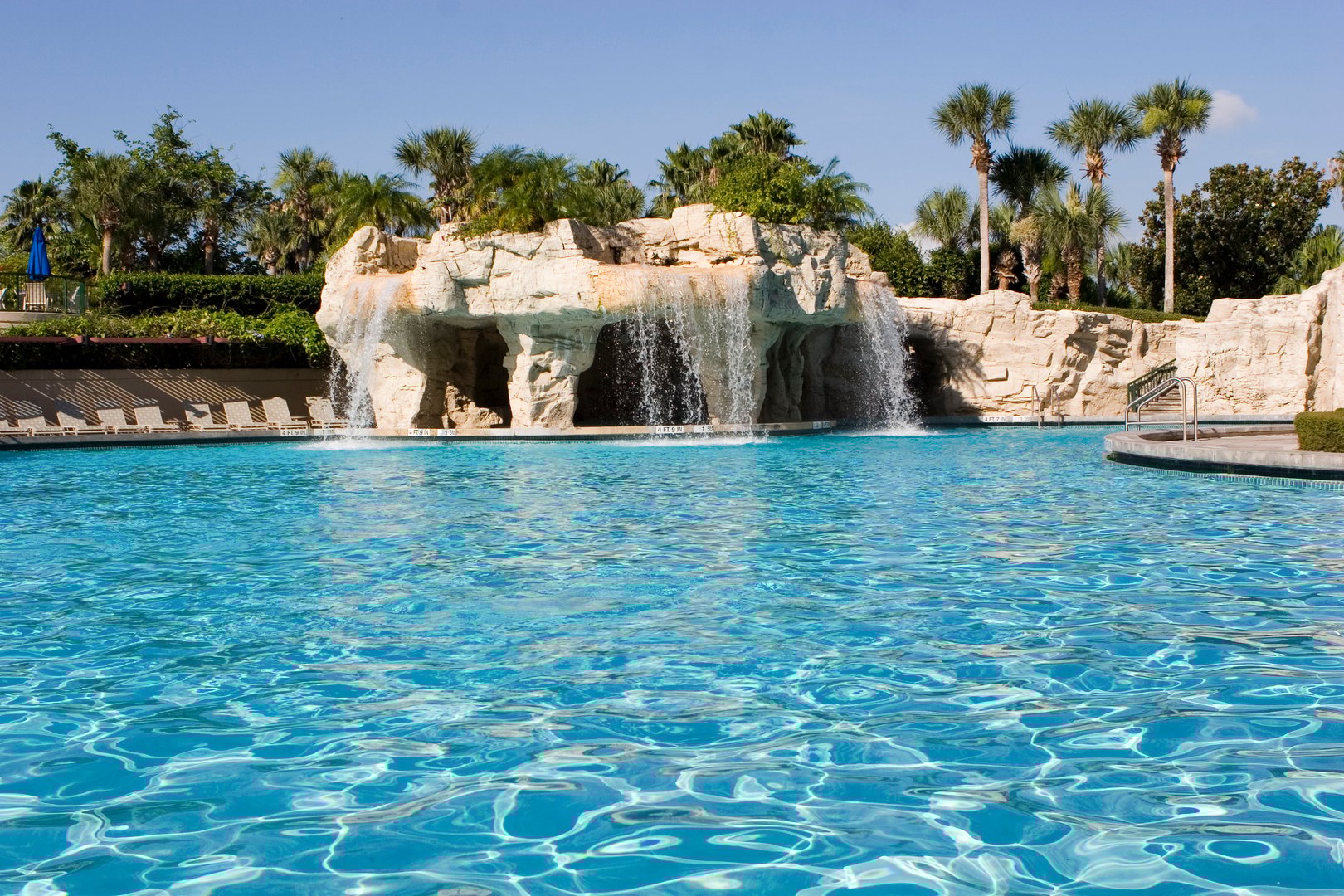A clear blue swimming pool with a stone waterfall feature and palm trees