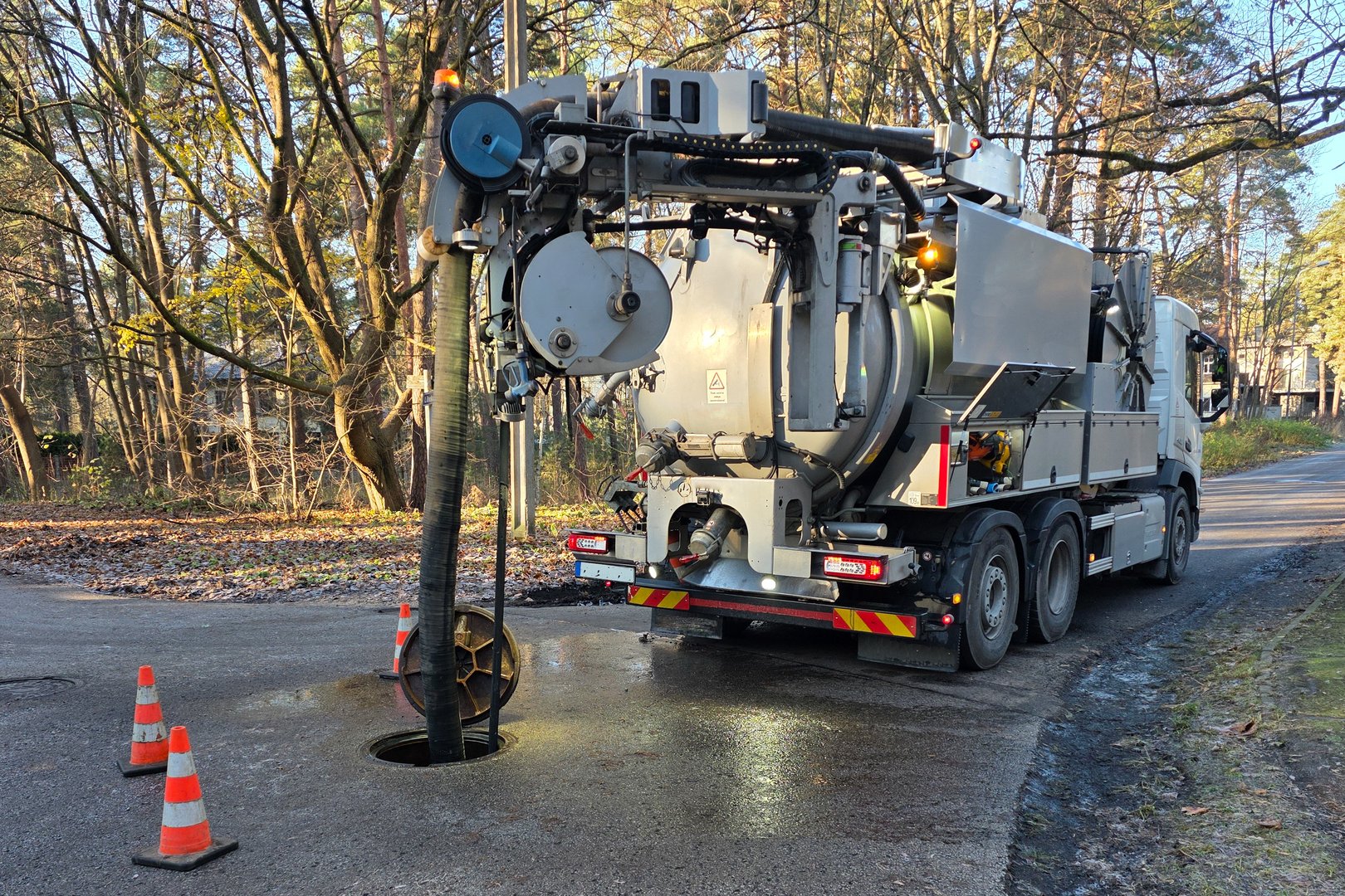 A hydro-vac sewer cleaning truck removes debris from an underground pipeline through an open manhole, with safety cones placed on a quiet forest road