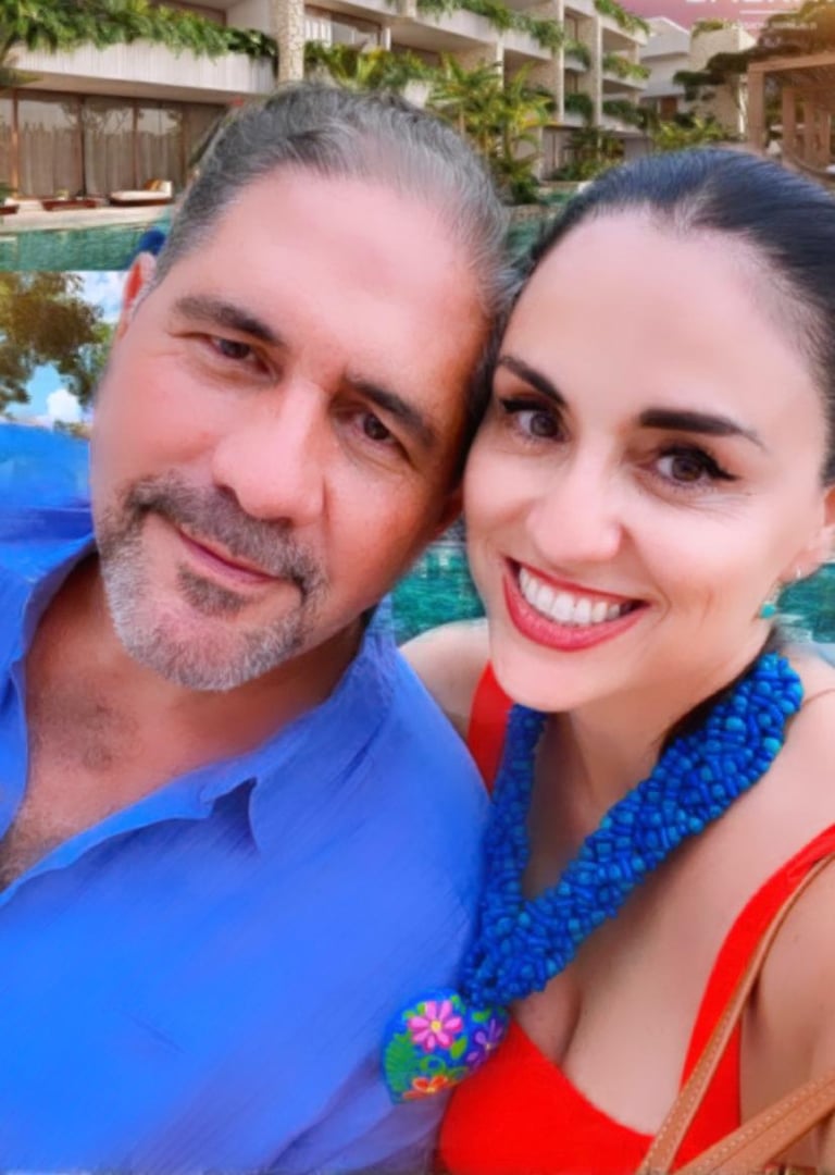 Smiling couple taking a selfie by a pool, man in blue shirt, woman in bright top with blue necklace.