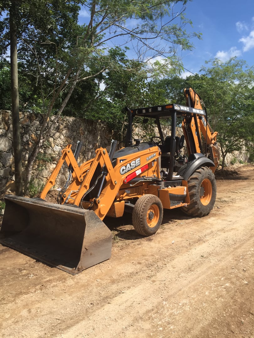 Orange Case backhoe loader parked on a dirt path with trees and a stone wall in the background.