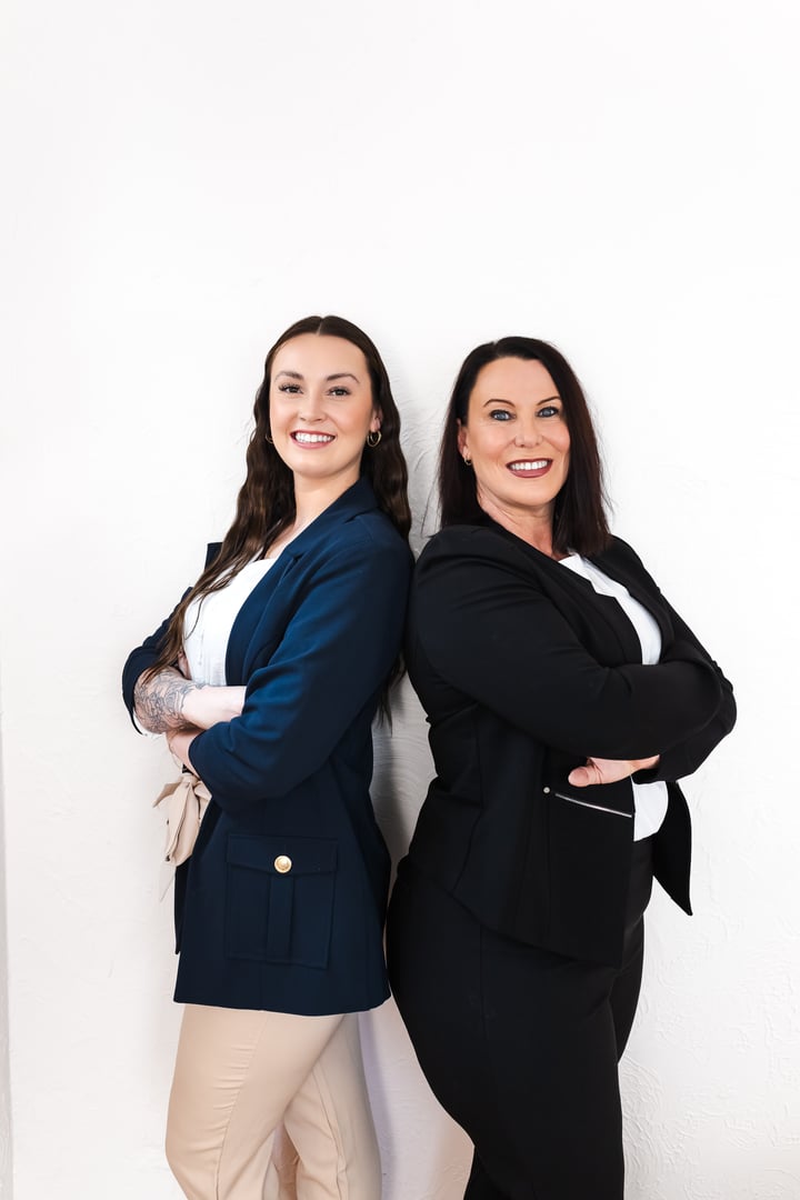 Two women in business attire standing back to back, smiling, against a white background.