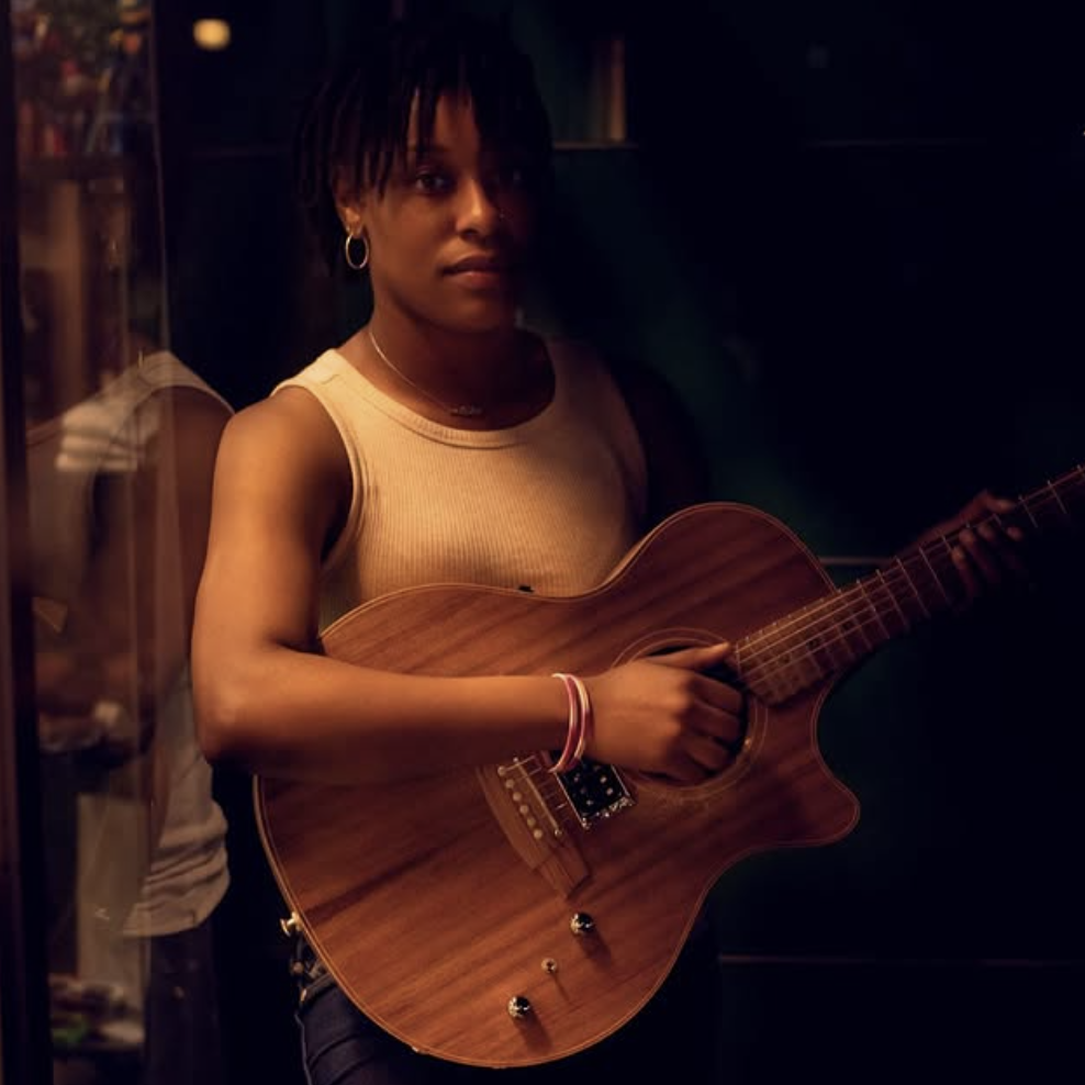 Person wearing a white tank top holds an acoustic guitar, standing against a dark background with one hand on the strings.
