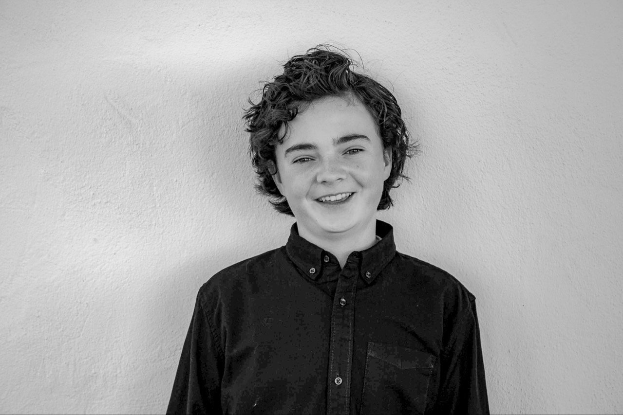 Black and white photo of a smiling boy with curly hair wearing a dark button-up shirt, standing against a plain wall.