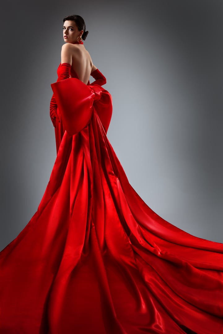 Fashion Woman in Red Silk Backless Dress with Oversized Bow side View. Elegant Lady in Open Back Satin Maxi Gown over Gray Background. Beautiful Model in Wedding Dress with Long Silk Train Fabric