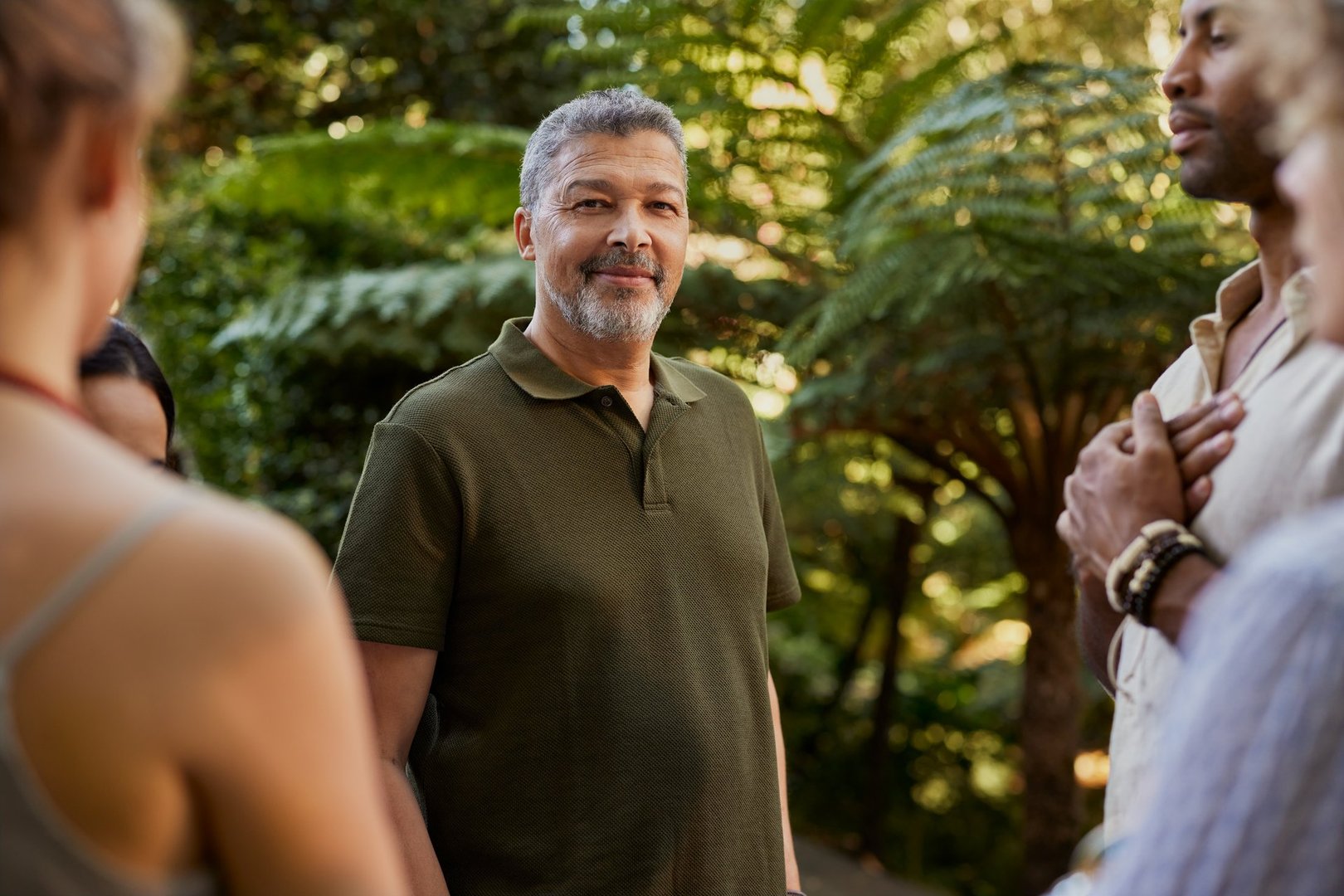 Mature man stands calmly at wellness retreat surrounded by nature and multiethnic people. Serene mixed race man in green shirt looking at camera peacefully during a mindful group gathering in the forest. Positive mature happy confident hispanic man smiling at camera.