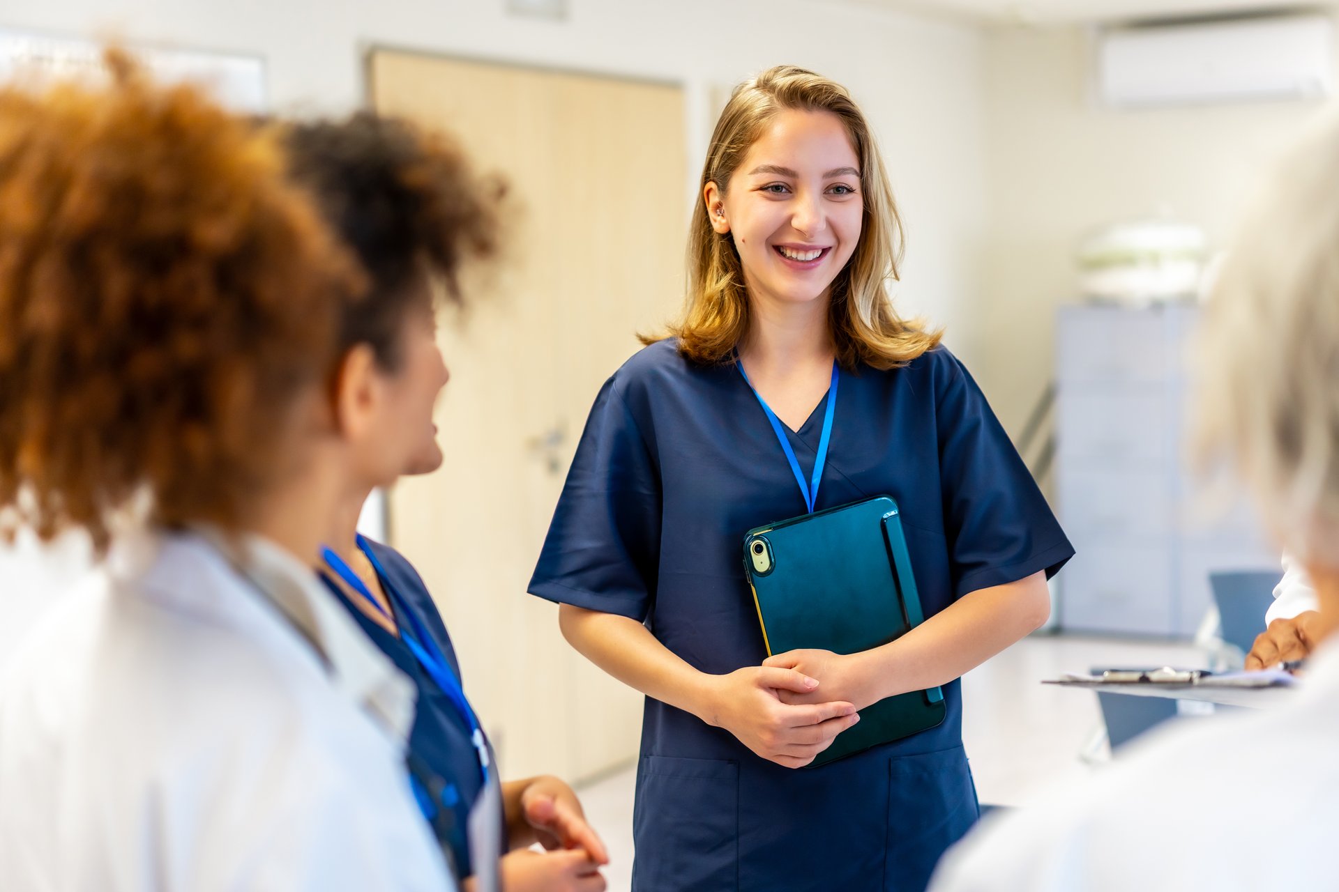 Young nurse smiling and engaging with colleagues while holding a tablet in a bright hospital hallway, fostering teamwork and communication