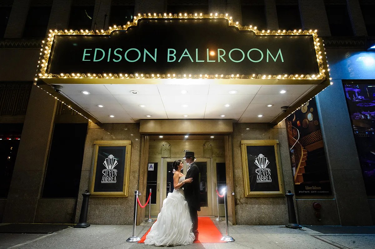A couple in formal attire poses on a red carpet under the illuminated sign of Edison Ballroom at night.