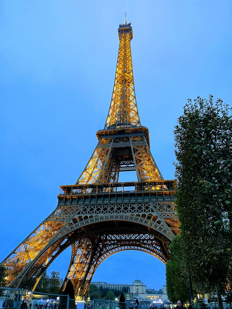 The Eiffel Tower illuminated at dusk, surrounded by trees and a clear blue sky, with people walking nearby.