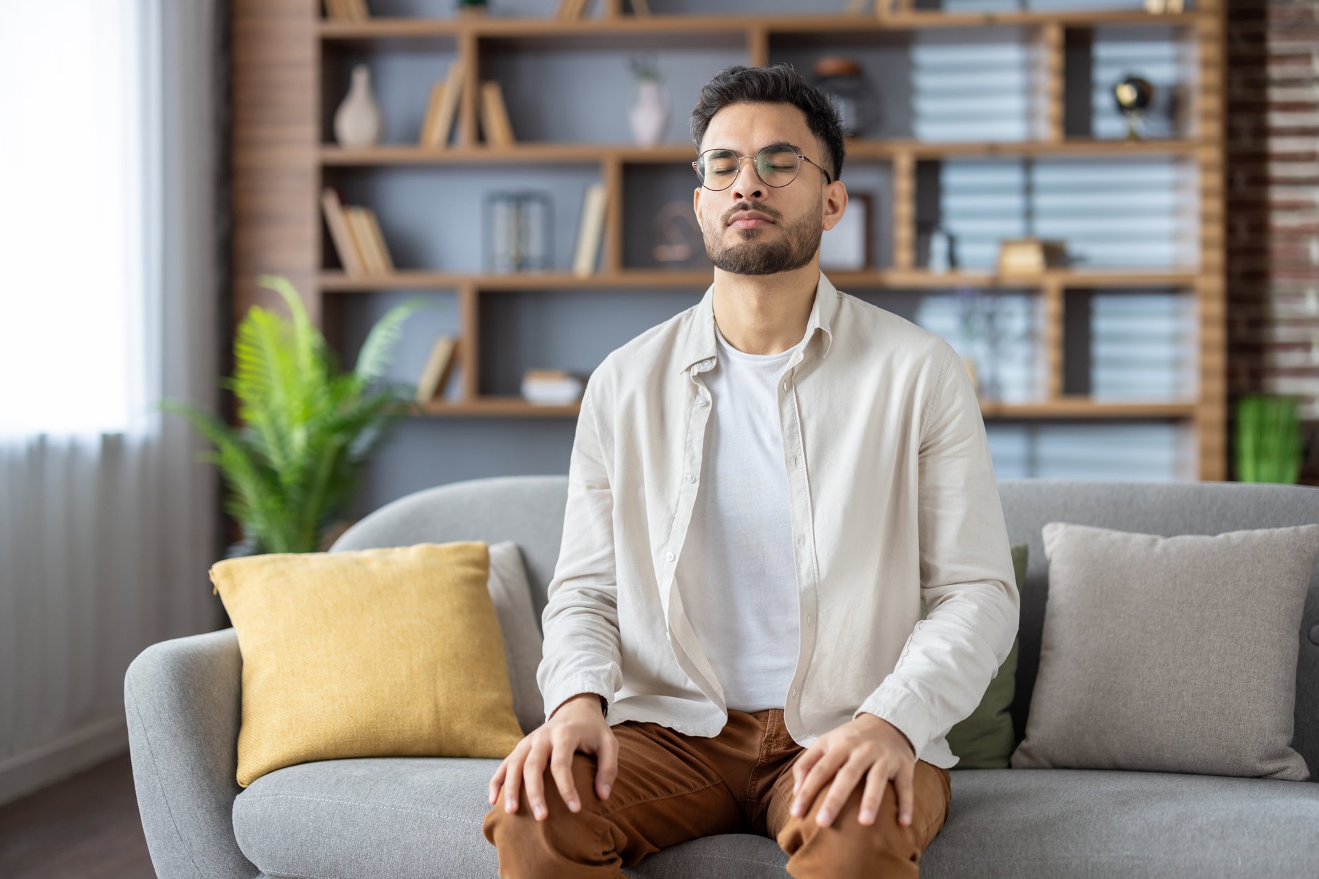 Young man sitting on sofa with eyes closed, practicing meditation and mindfulness. He is focusing on relaxation, inner peace, and mental clarity in cozy home setting.