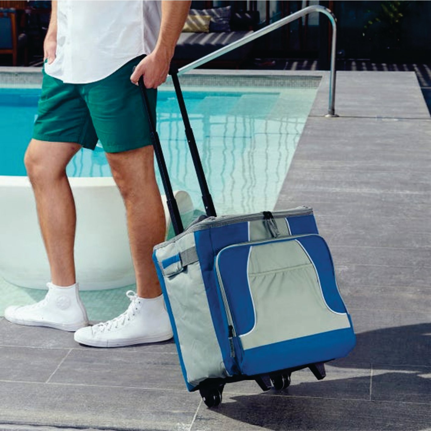 Person in shorts and sneakers pulling a wheeled cooler bag by a pool.