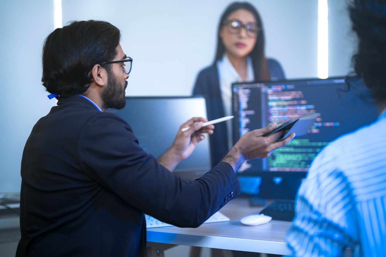 Diverse team of AI developers collaborating in a modern office. Indian male leading a discussion with Asian and Hispanic female colleagues on a machine learning project.