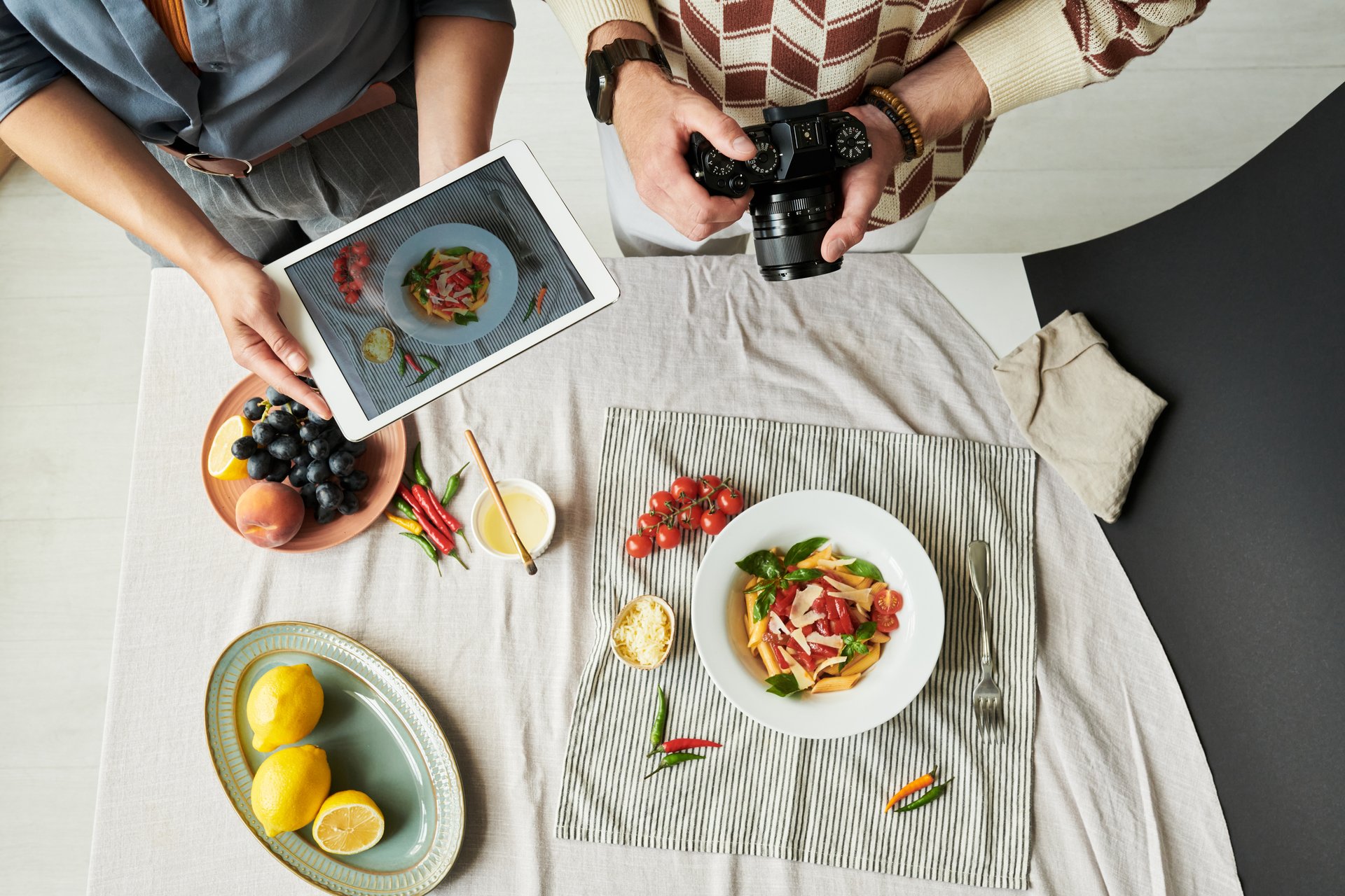 Woman holding tablet photographing salad while man using camera capturing food on table with fruits, vegetables, and copy space