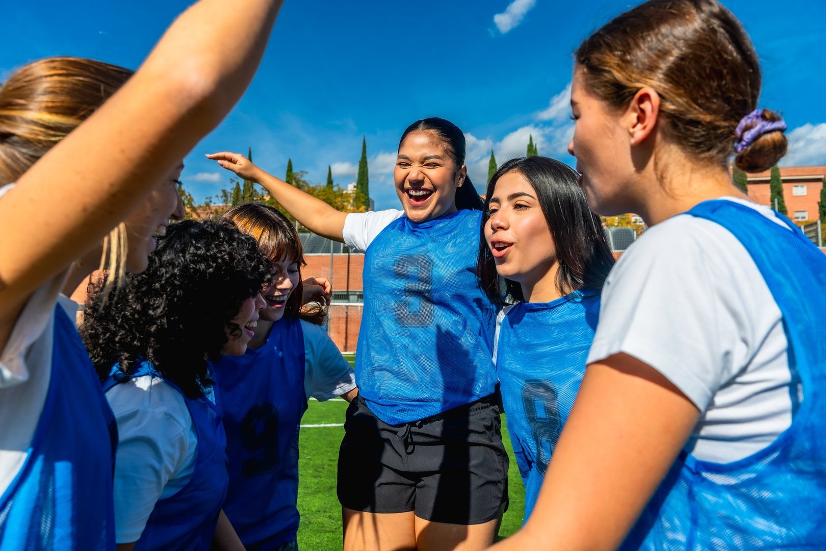 Group of excited young adult women soccer players huddling together and celebrating a victory on an outdoor sports field under a clear blue sky, showing teamwork and joy