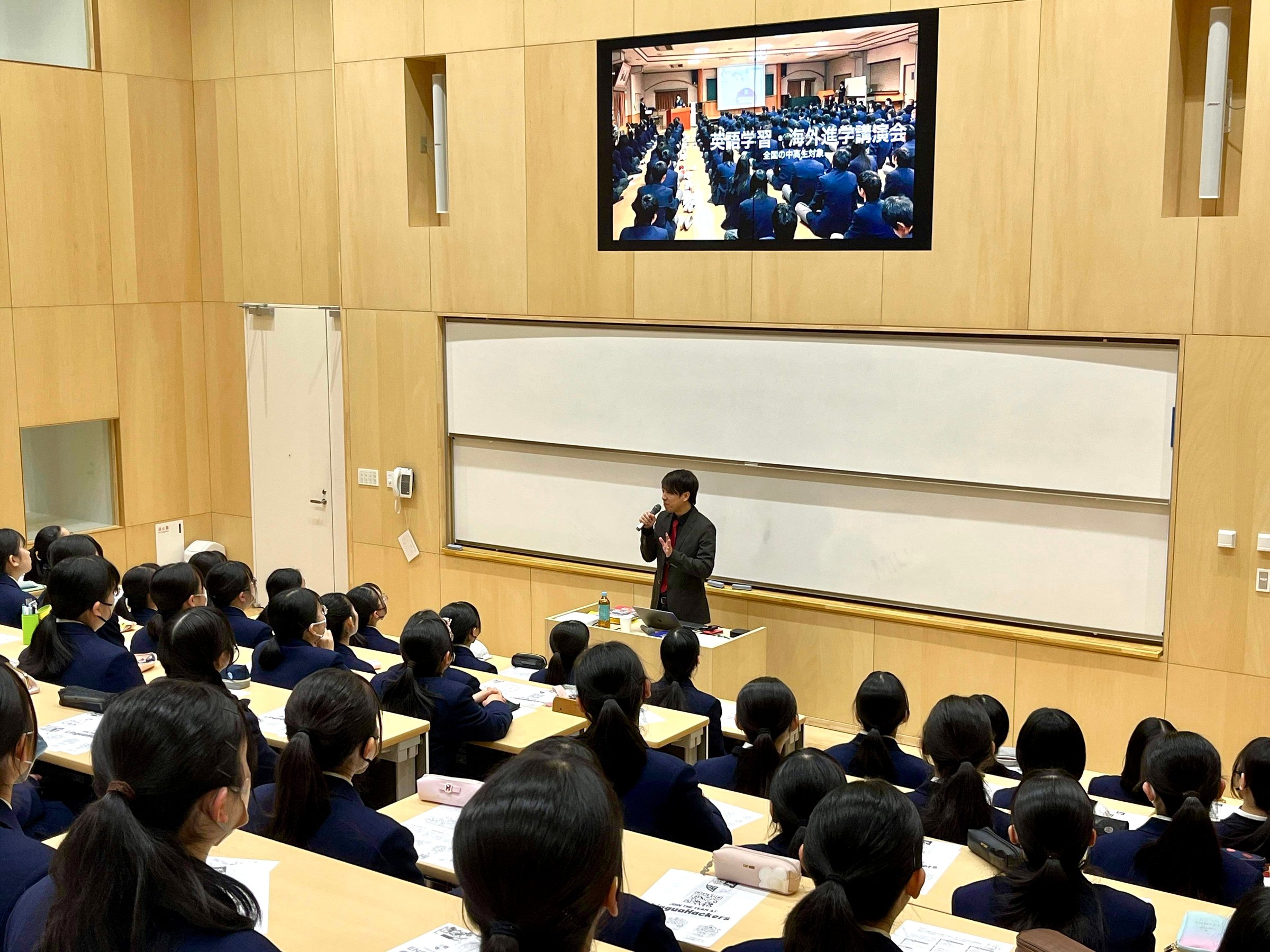 A speaker in a lecture hall addresses students in uniforms, with a large screen displaying a related image above.