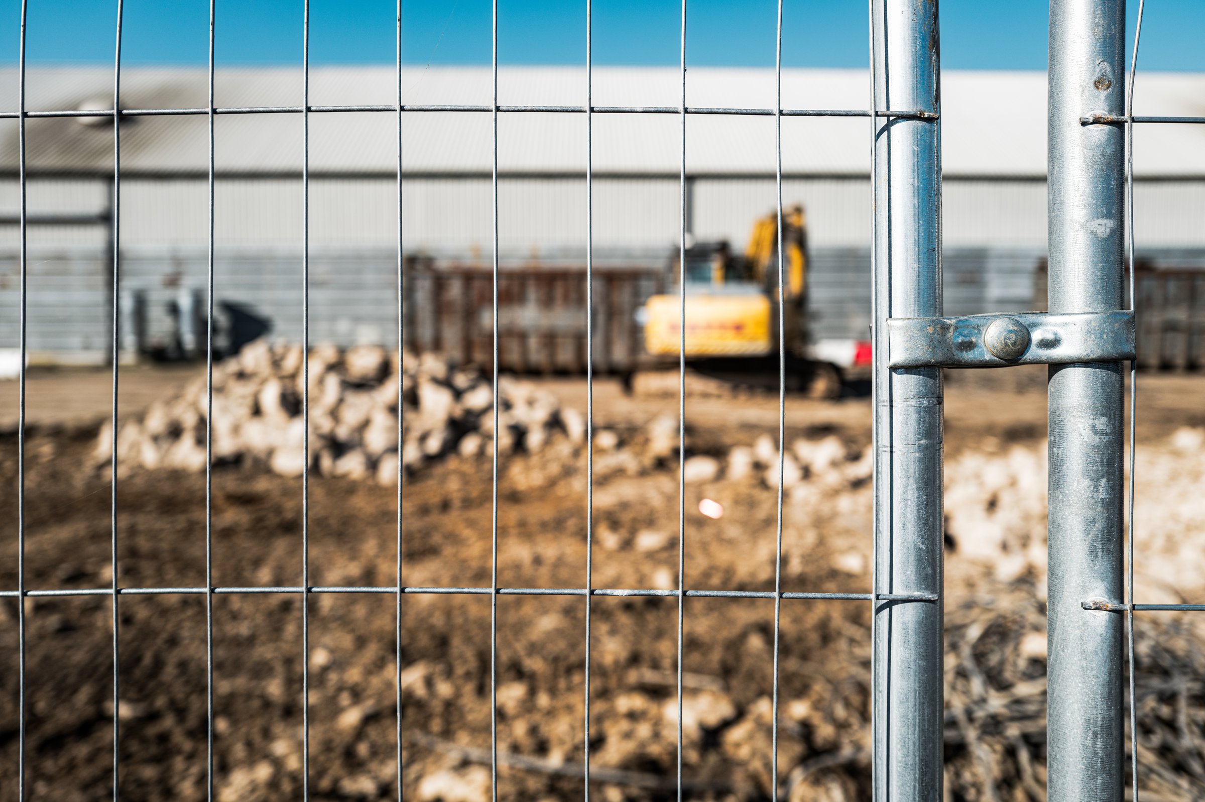 Shallow focus of a secure metal fence seen in the vicinity of construction works on a British industrial estate. A large yellow excavator can be seen near the rubble.