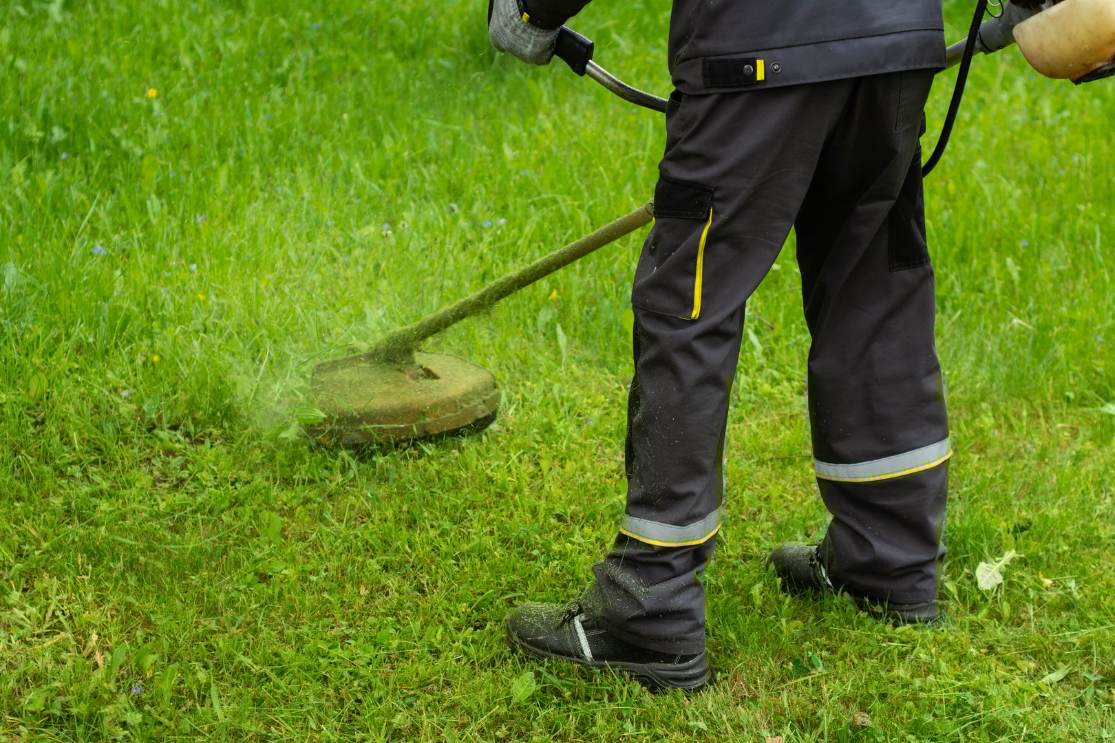 Worker in uniform mowing grass with a string trimmer in a public green area. Manual labor and professional landscaping work