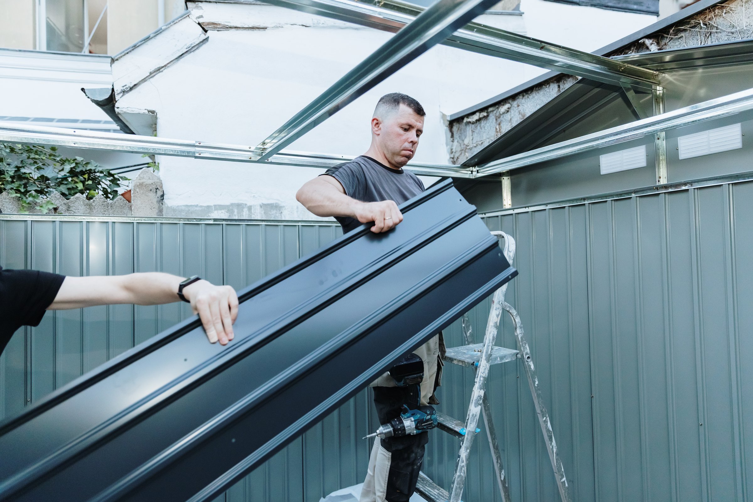 One Caucasian young worker standing on a stepladder and taking steel roofing sheets from his assistant to install on a metal shed in the backyard on a summer day, side view, medium shot. Concept assembly metal shed, time teamwork, using technology.