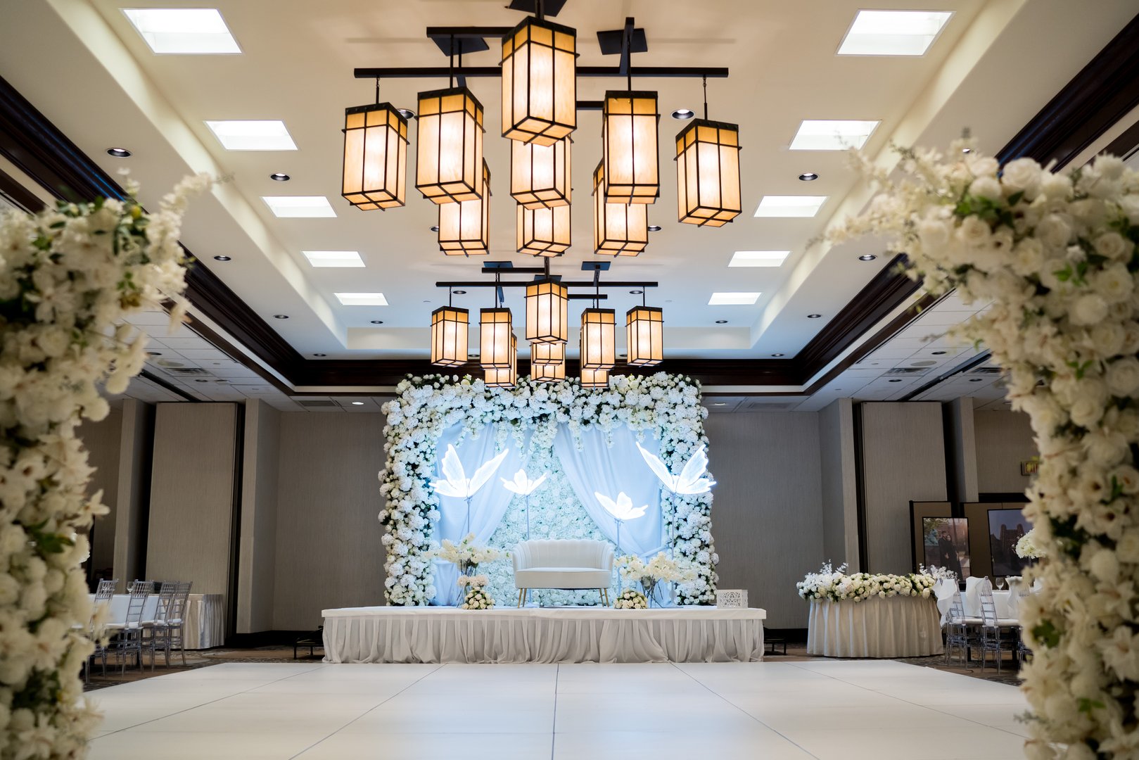 Elegant wedding venue with white floral arch, stage with backdrop of flowers and fabric, and warm hanging lanterns.
