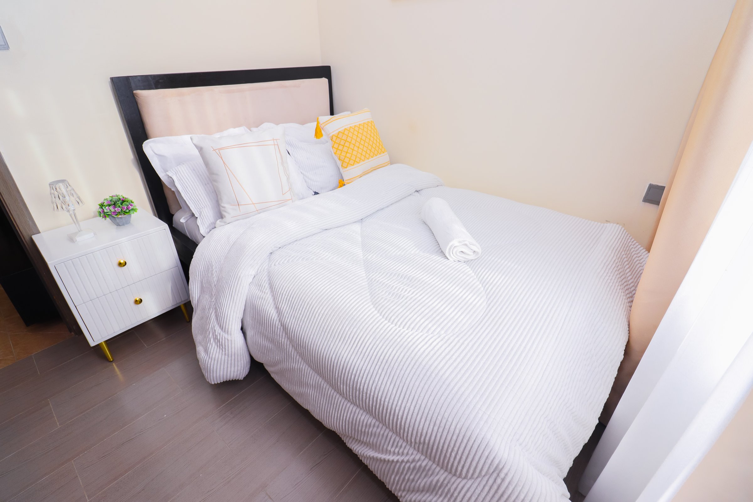 A neatly made bed with white bedding, decorative pillows, and a rolled towel on top. A nightstand with a lamp and plant is beside it.