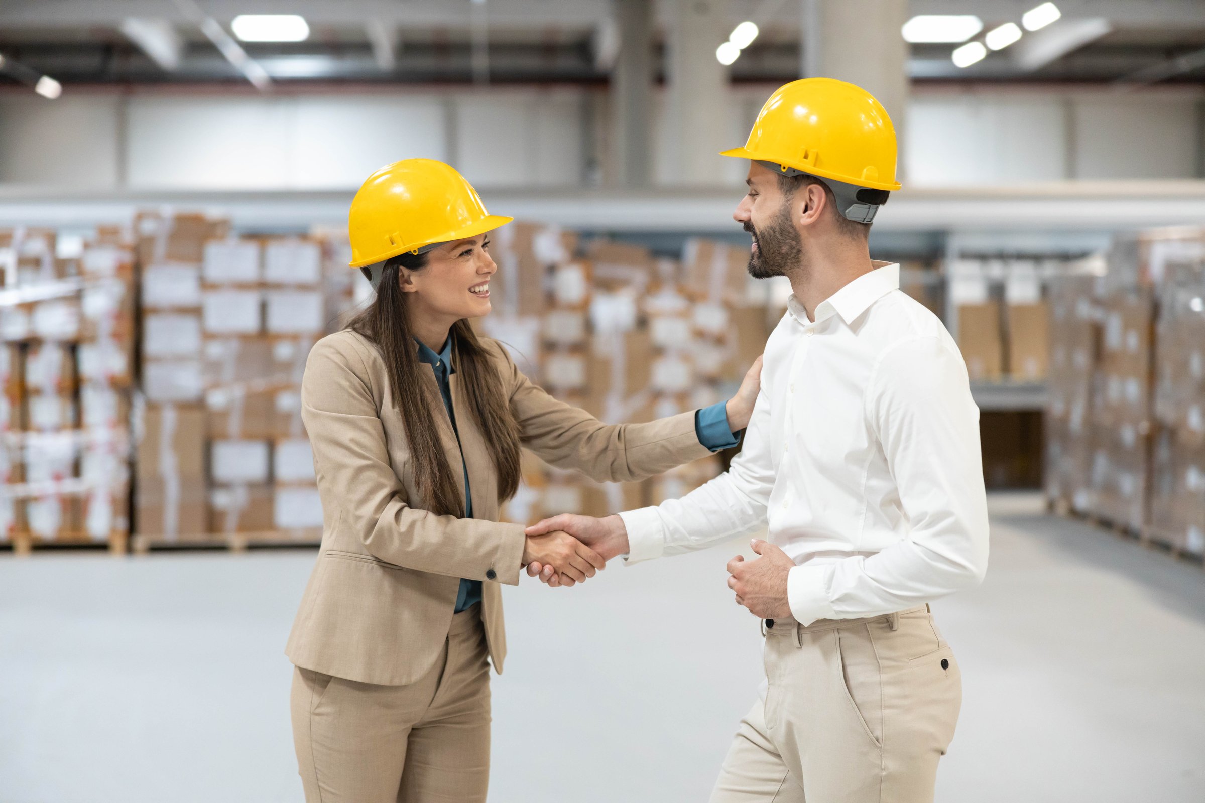 Businesswoman and businessman shaking hands while wearing safety helmets in a warehouse. Agreement and trust in logistics management.