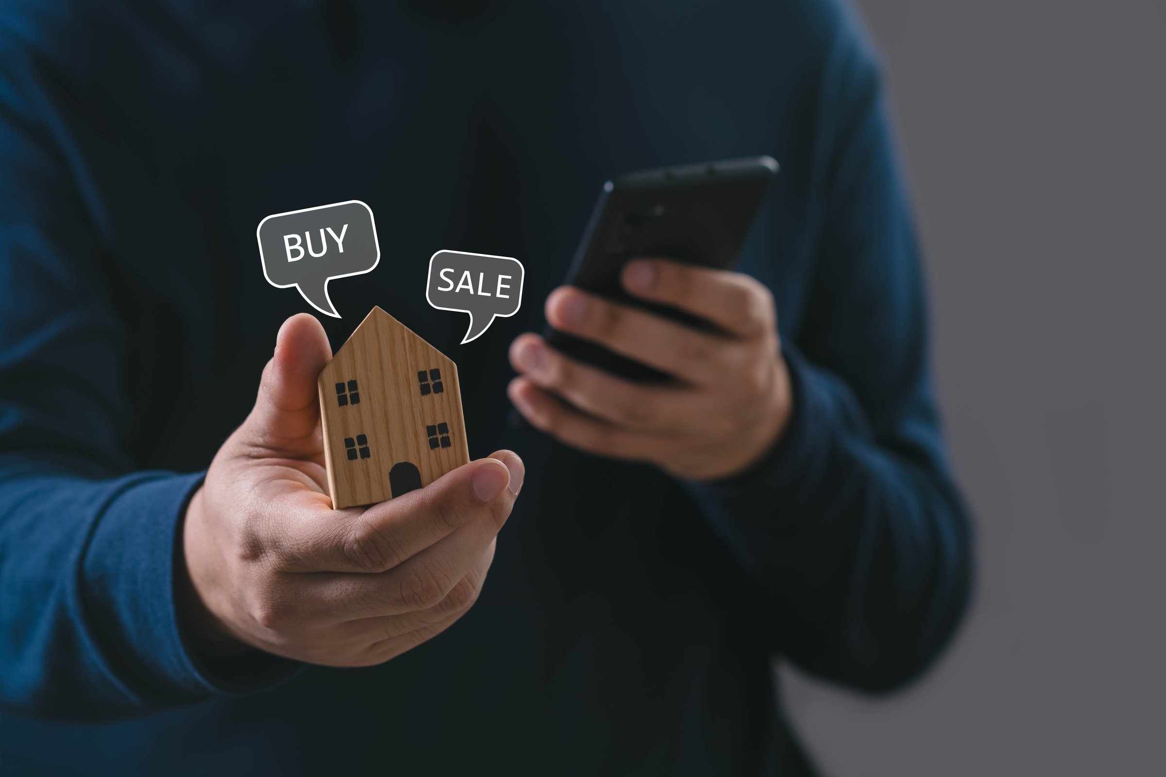 holding wooden house model with buy and sale icons while using smartphone, representing real estate transaction, online property market, home investment, digital real estate service and mobile buying.