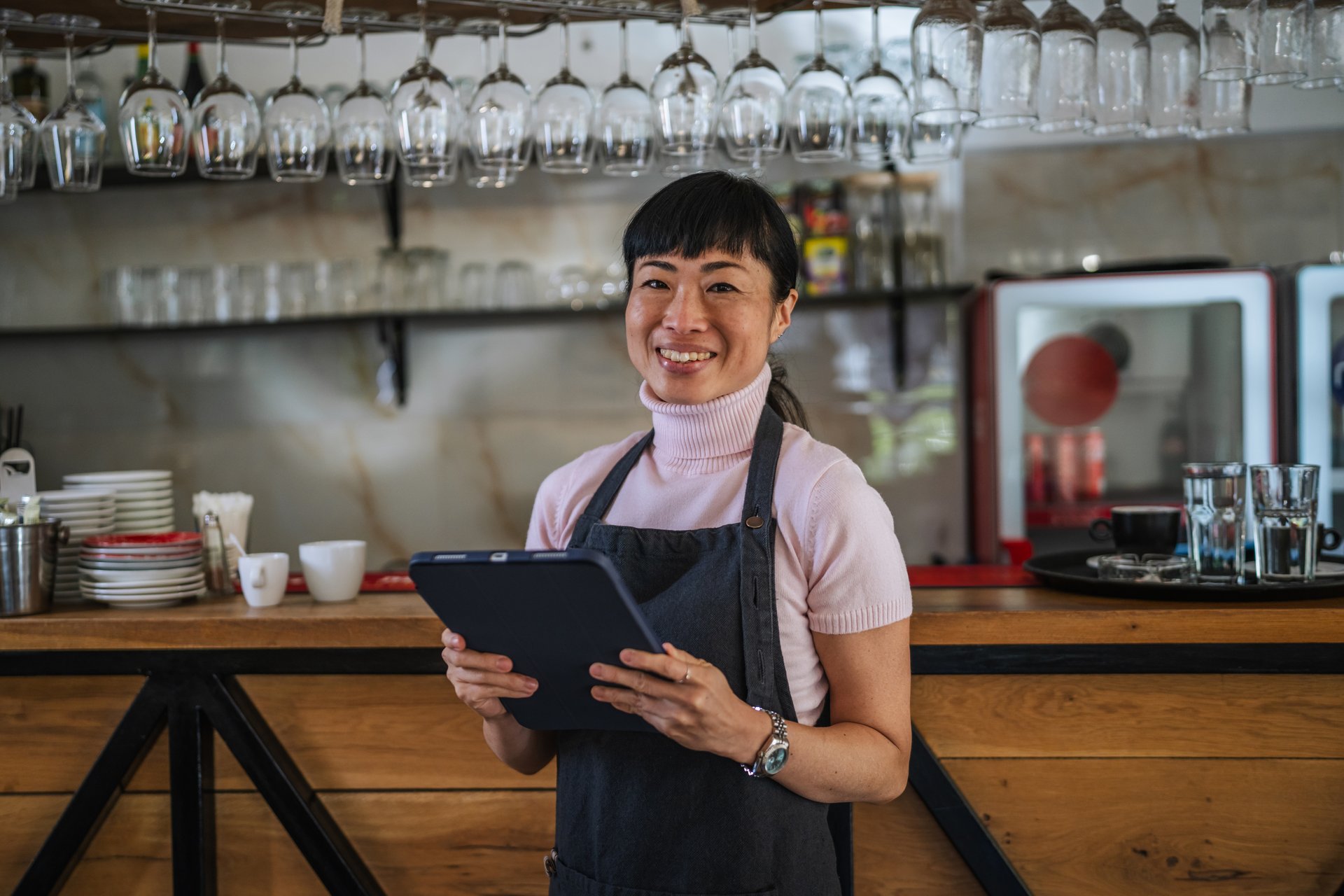 Asian woman cafe owner smiling confidently behind the counter, holding a tablet and using modern technology to manage orders, inventory and daily small business operations