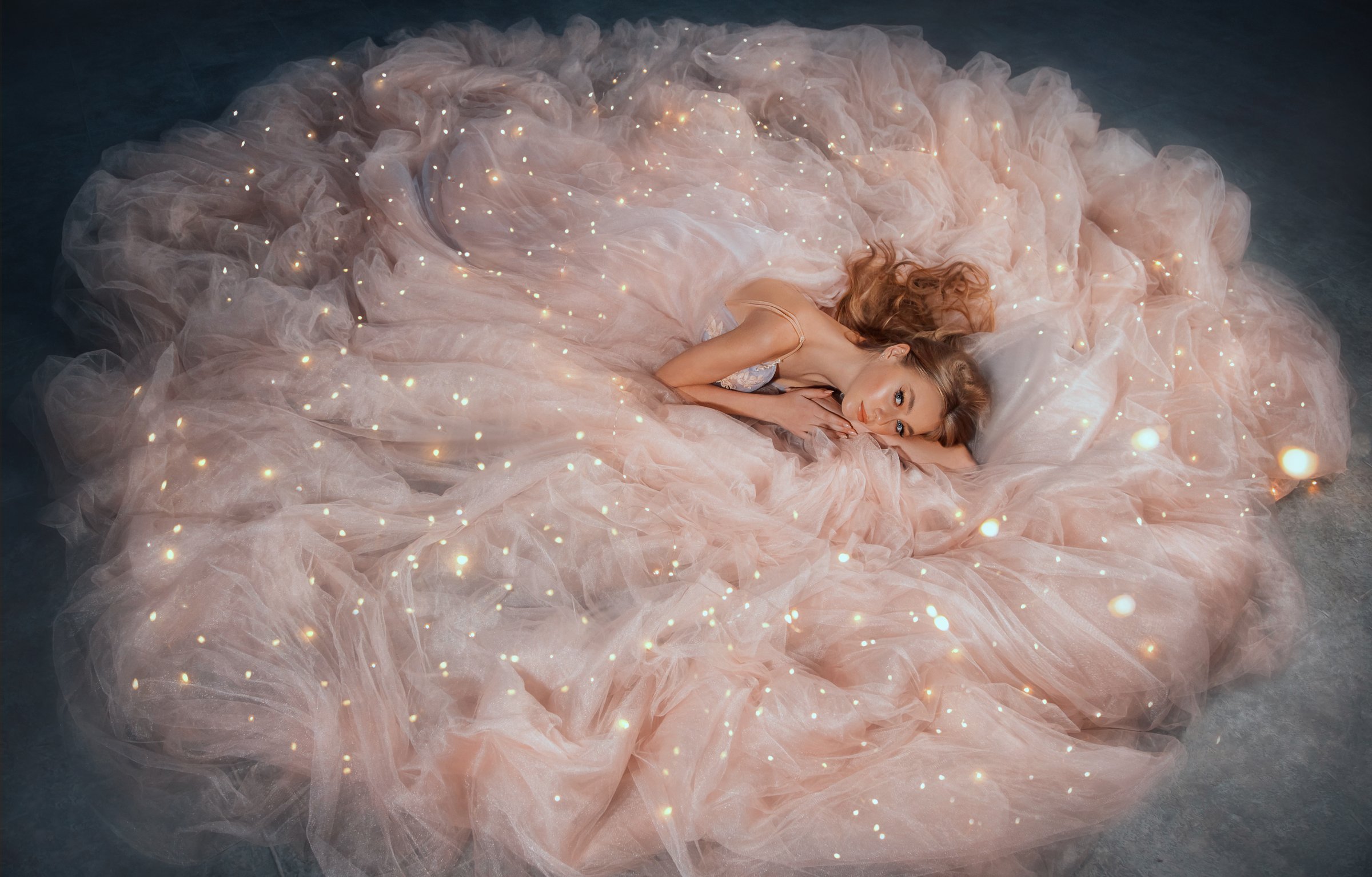 Fairy queen fashion model in luxurious shining pink dress posing in studio. Princess girl, peach outfit with sequins lies in fabrics of outfit, long skirt gown. Fantasy woman sleeping beauty woke up