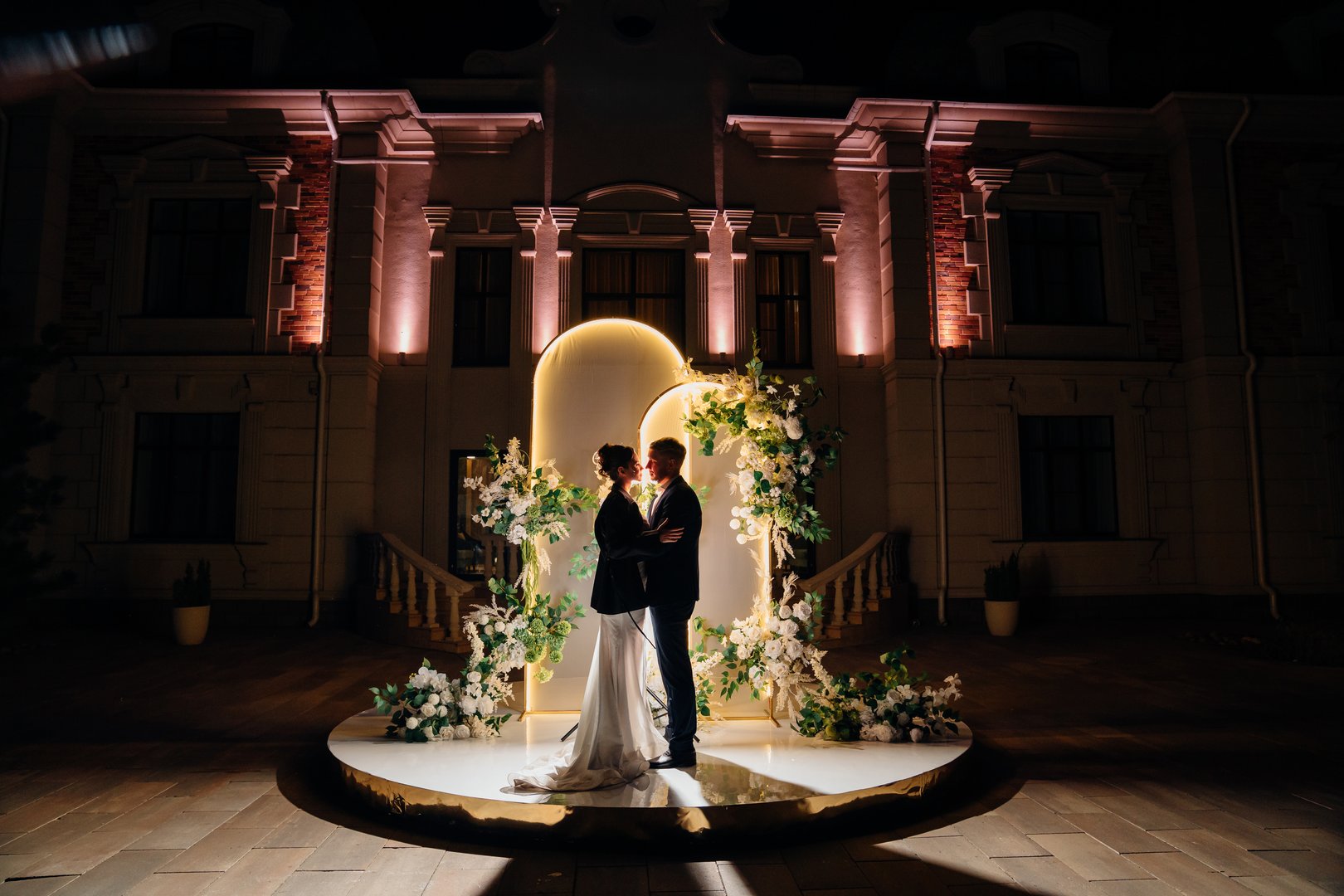 Silhouettes of a bride and groom in front of a wedding arch decorated with flowers and greenery, illuminated. The evening setting creates a romantic atmosphere. softly lit building in the background.