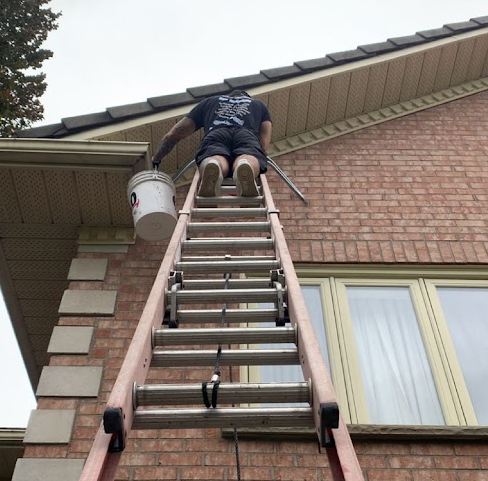Person on ladder near roof, holding a paint bucket, doing maintenance on a brick house with beige trim and shingles.