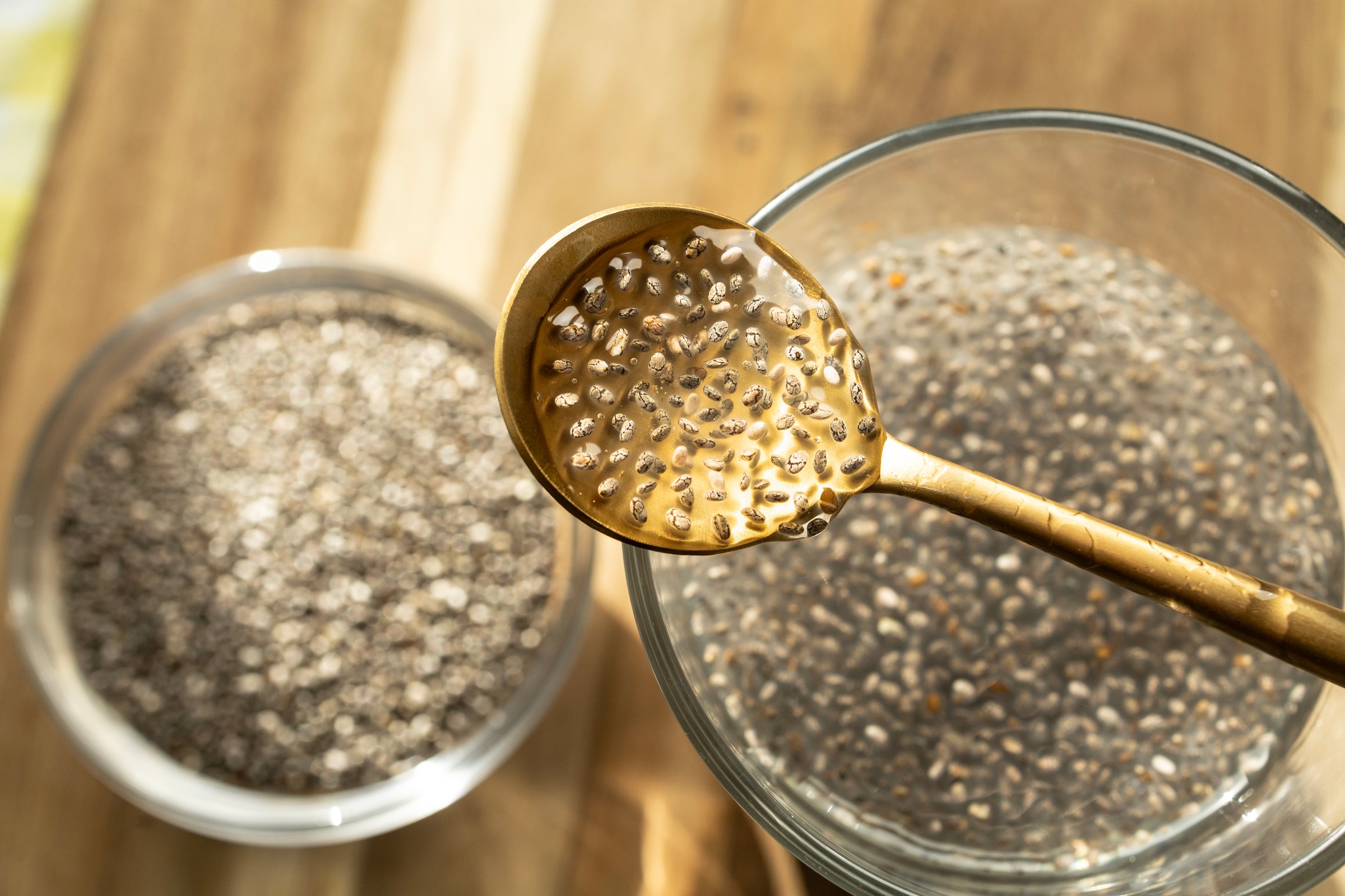 A warm and natural scene showing chia seeds in a glass of water with a golden spoon. The seeds are releasing their gel-like mucilage, highlighting their transformation into a nutrient-rich superfood.