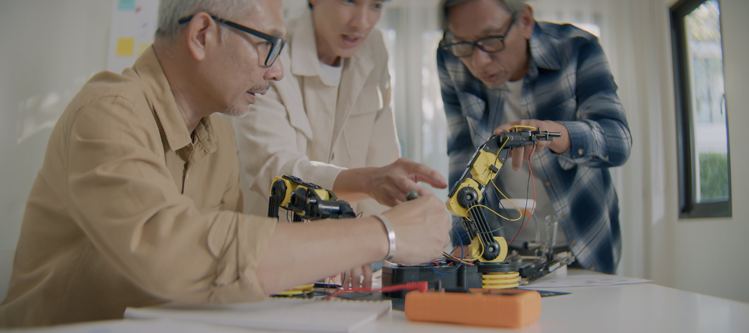 Group of Asian male engineers testing and assembling robotic arm prototype on table. Concept of robotics, engineering collaboration, and modern automation.
