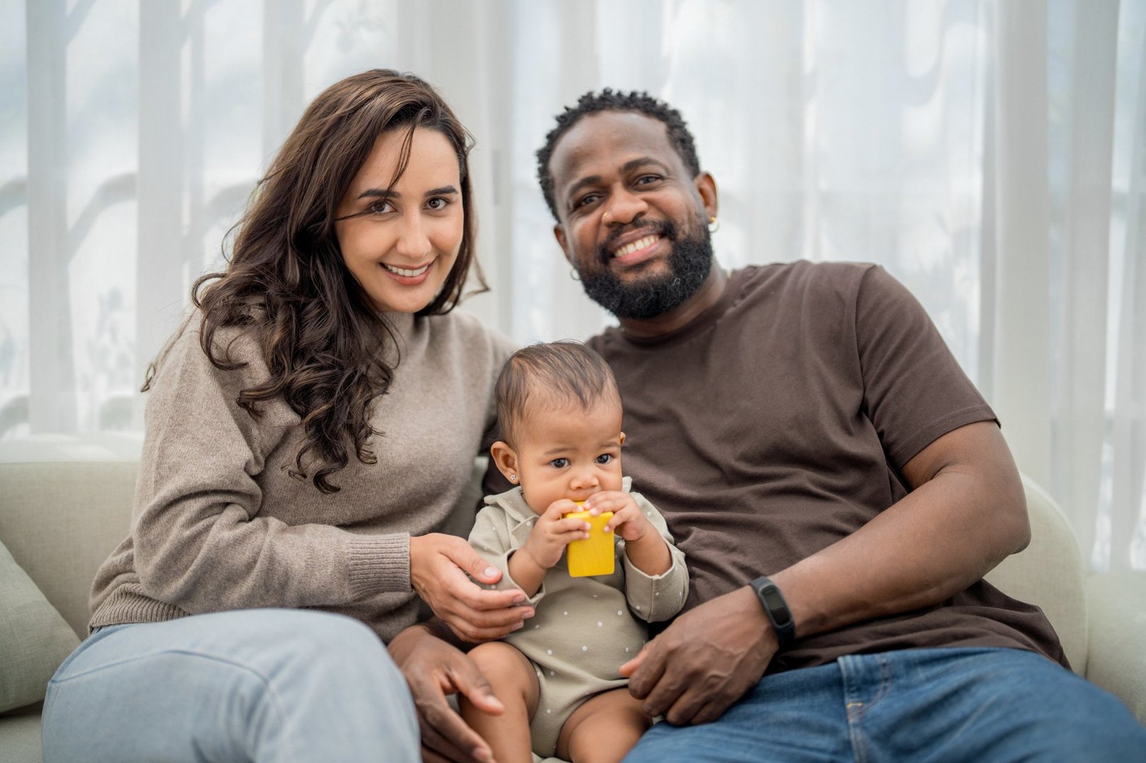 A happy family sits on a couch in their living room. The woman smiles while holding her baby, who is playing with a yellow toy. The man sits beside them, radiating joy in a warm atmosphere.