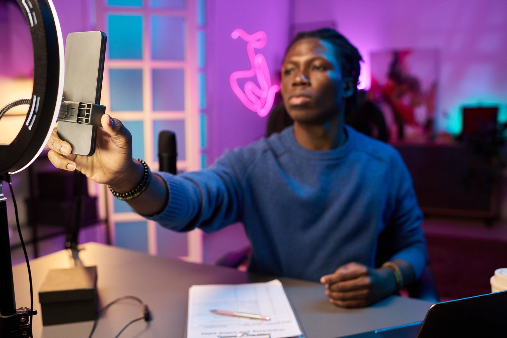 Young Black male vlogger adjusting smartphone on a ring light stand in a stylish home studio with ambient lighting. Desk featuring creative tools and notes in foreground