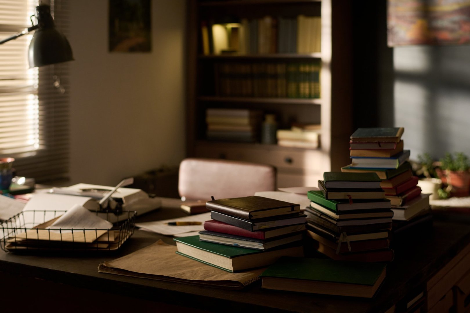 Stack of assorted books arranged on desk in home office with sunlight streaming through window, bookshelf filled with volumes in background, workspace suggesting academic or literary setting