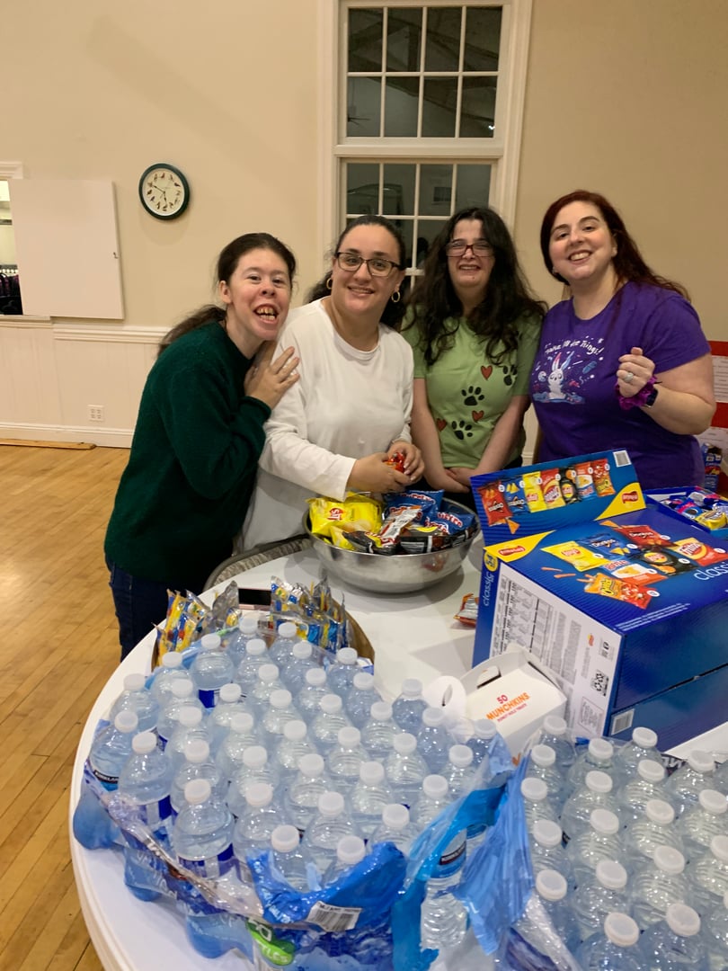 Four women smiling while preparing snacks and drinks on a table in a community hall.