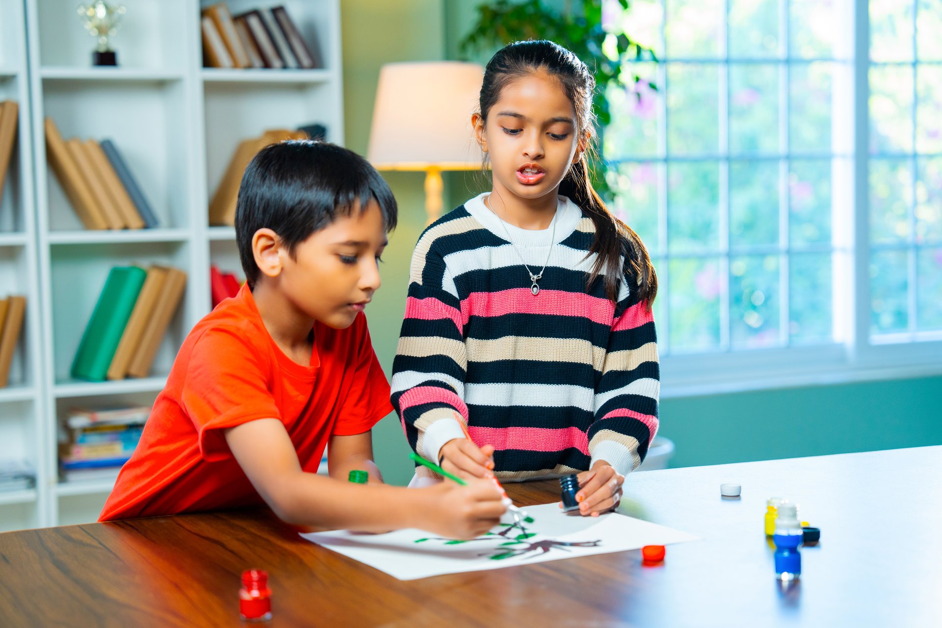 Indian Asian Cute Little Girl and Boy Siblings Playfully Painting on drawing paper Using Brushes at Home, Exploring Creativity and Art Skills, Sitting Together at Table in modern home
