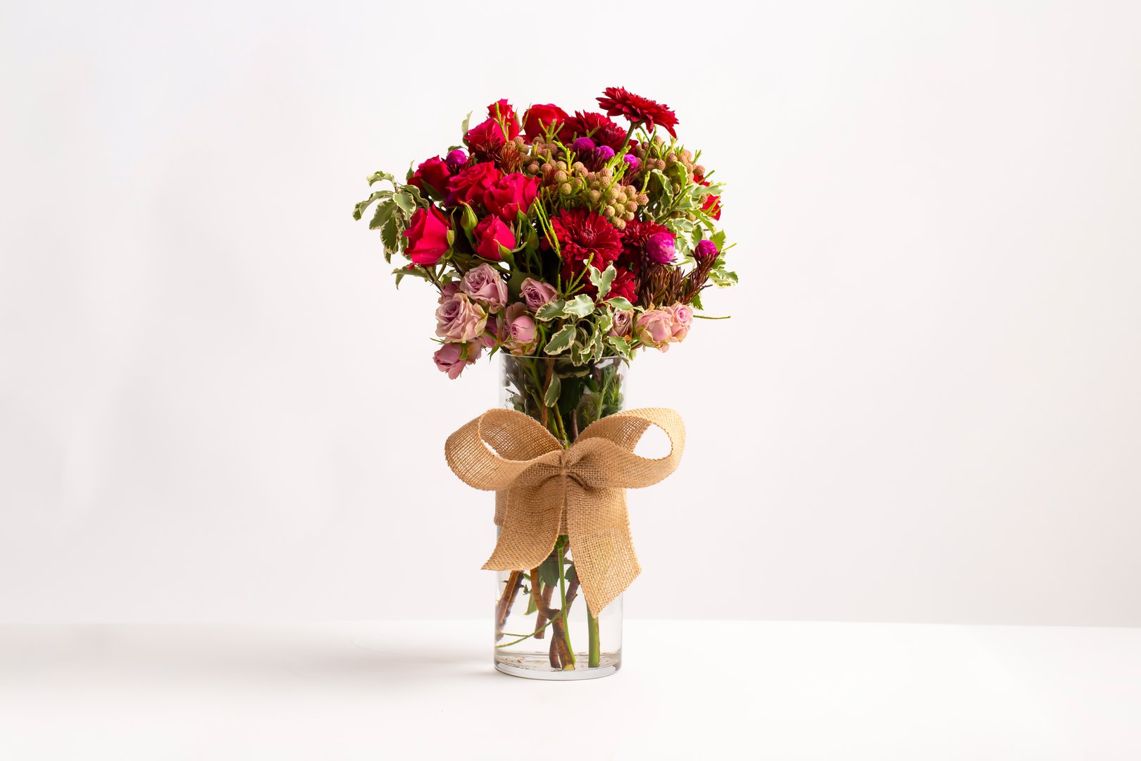 Rustic Floral Arrangement in Vase A vibrant bouquet of red roses, chrysanthemums, and other seasonal blooms arranged in a tall glass vase tied with a rustic burlap bow on white background side view